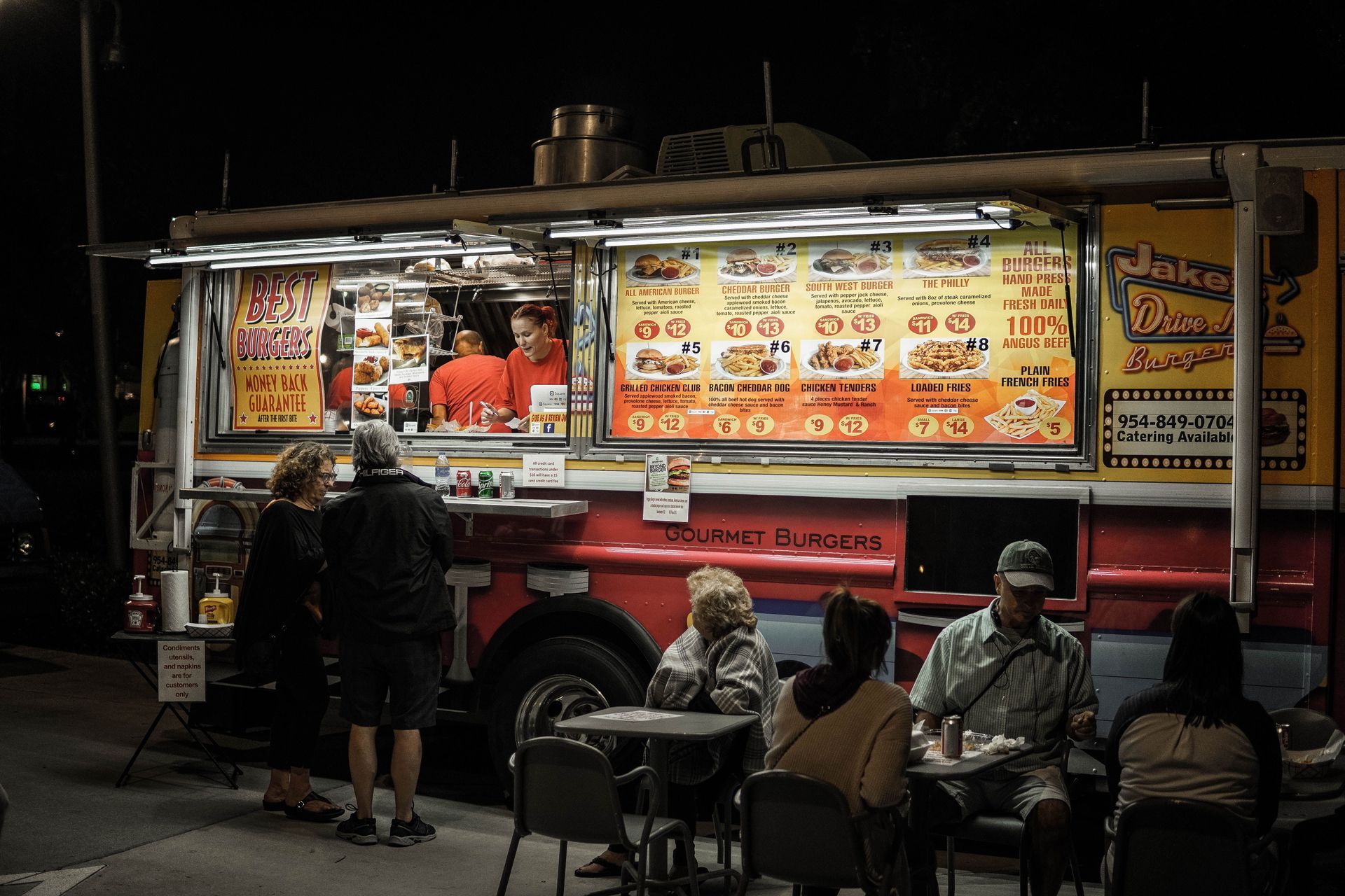 Night food truck with illuminated menu and people seated at tables waiting to order