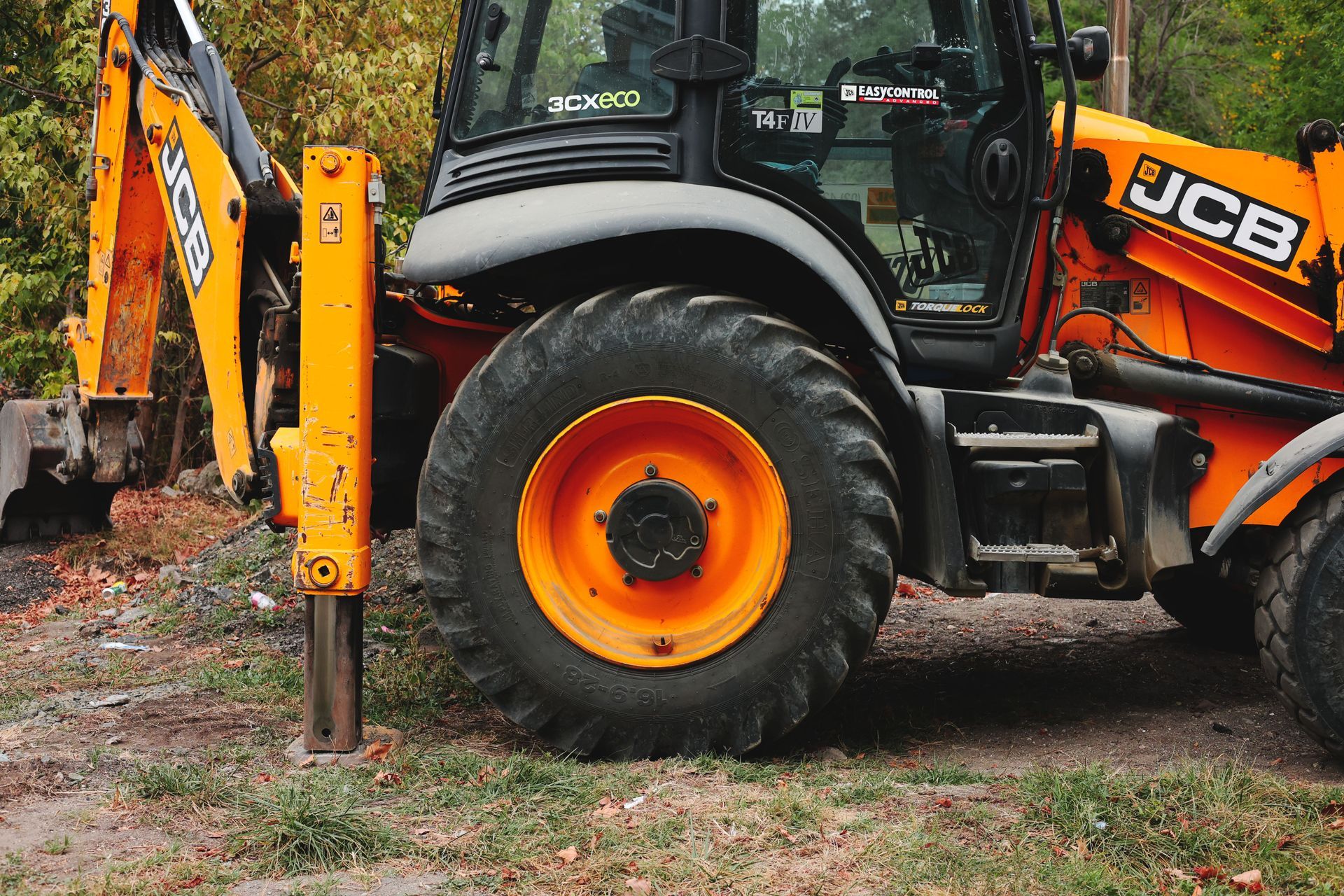 JCB backhoe loader with orange arm and large rear wheel on muddy ground, excavating near a tree