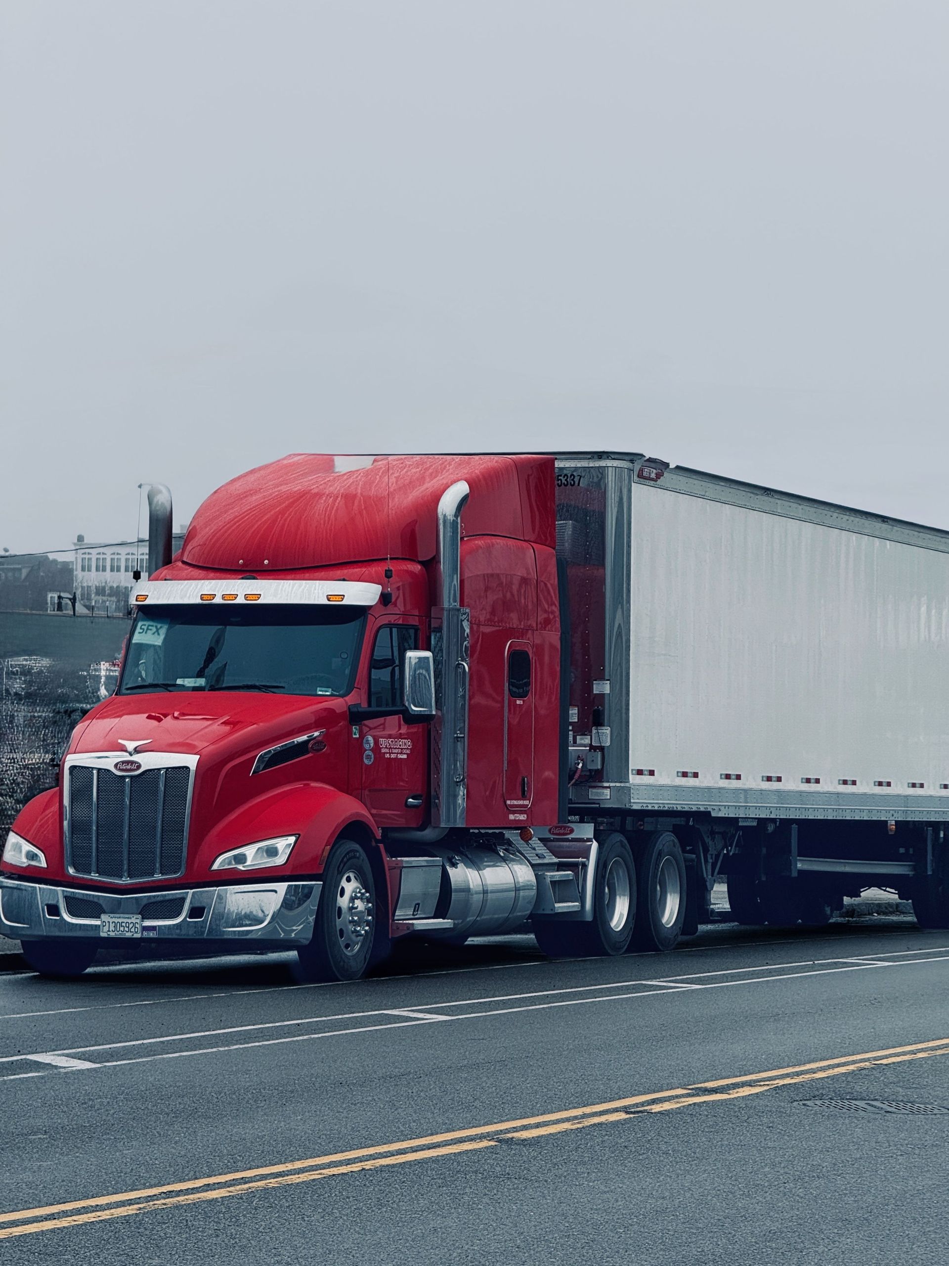 Red semi-truck hauling a white trailer on a wet road under an overcast sky