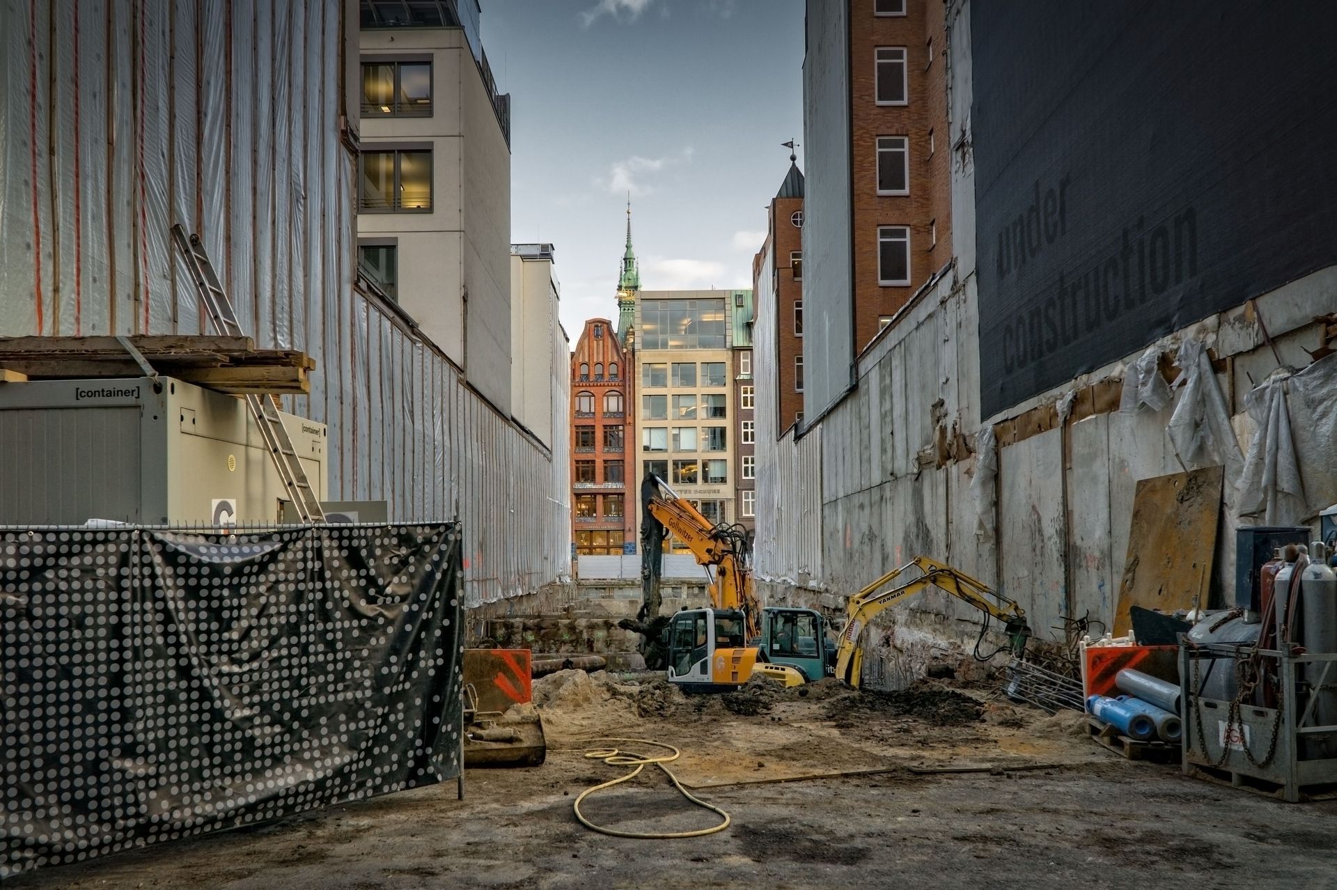 Construction site between tall buildings with excavators and debris along a narrow alleyway