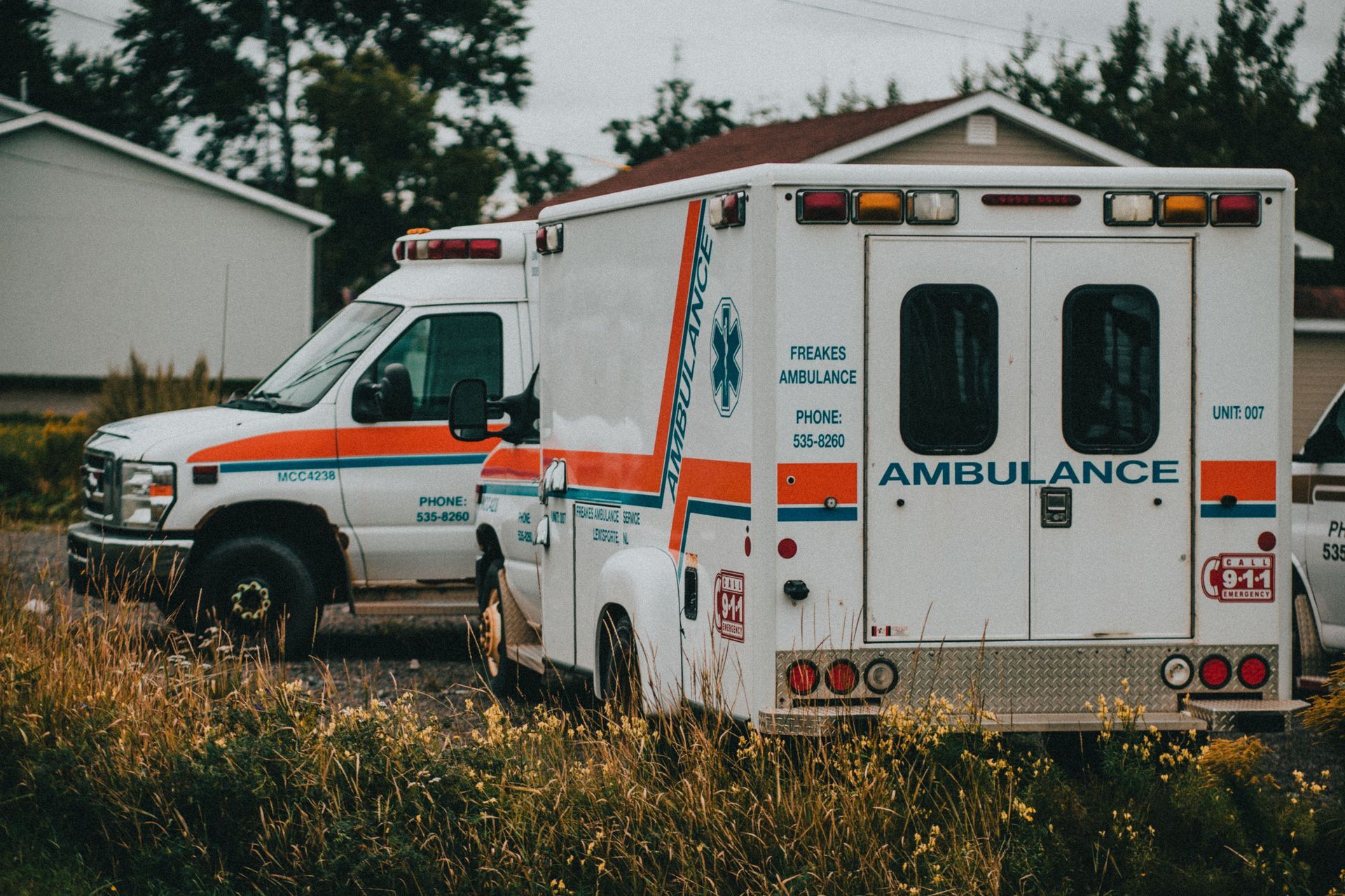 Two ambulances parked in tall grass outside houses, one showing “AMBULANCE” on the back