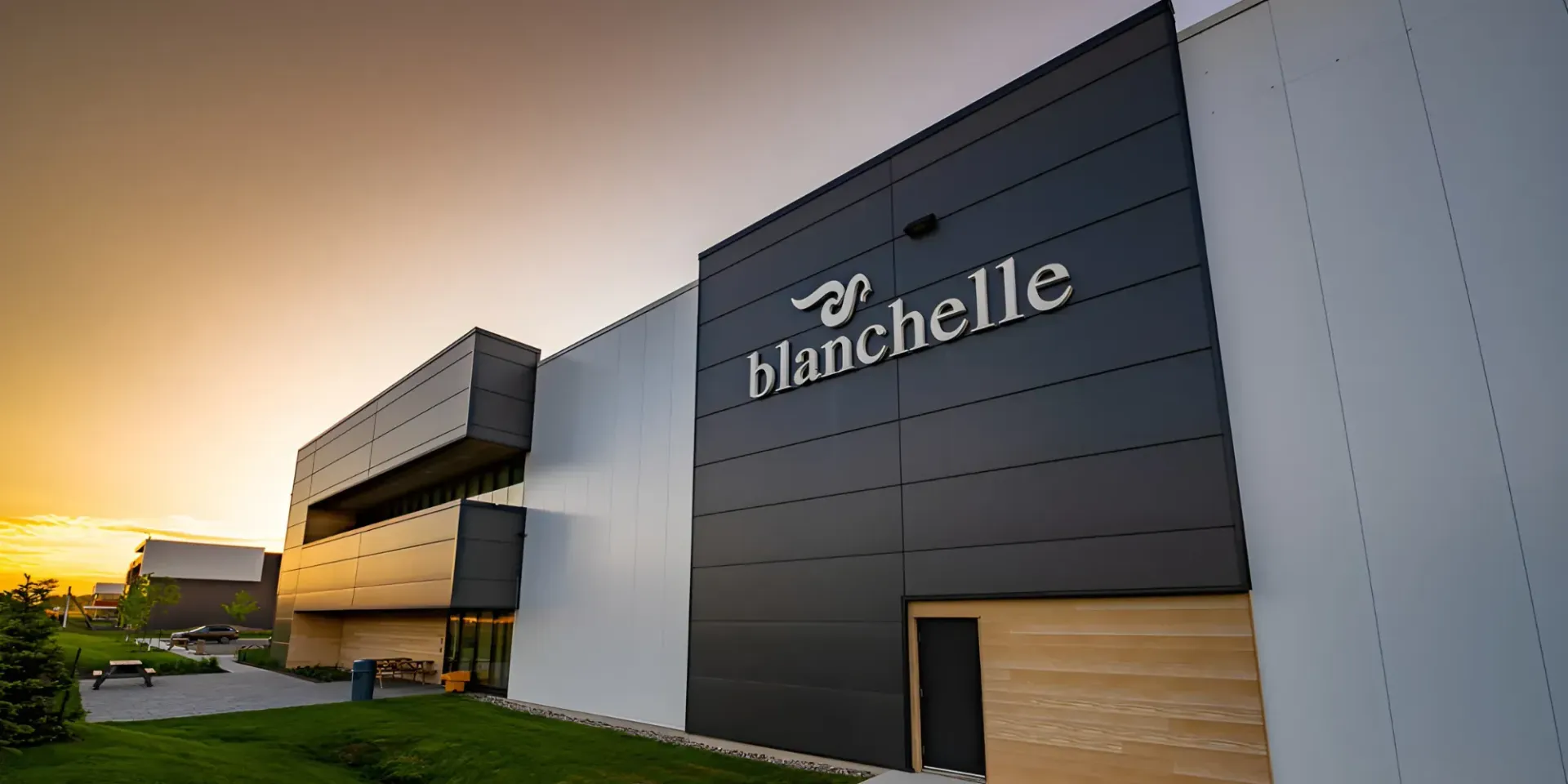 Bâtiment moderne à la façade sombre arborant le logo blanc « blanchelle » au coucher du soleil sur un ciel clair et doré.