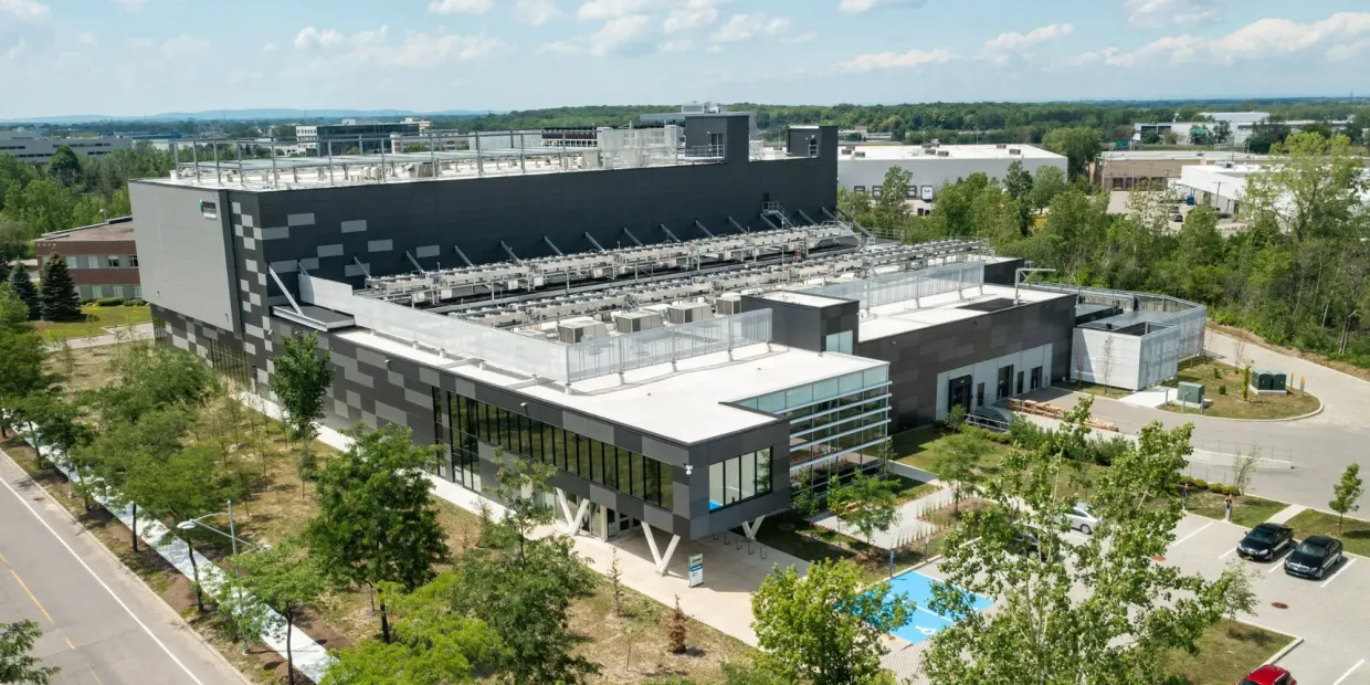 Vue aérienne d'un bâtiment commercial moderne gris avec des unités de climatisation sur le toit, entouré d'arbres, d'un parking et de routes.