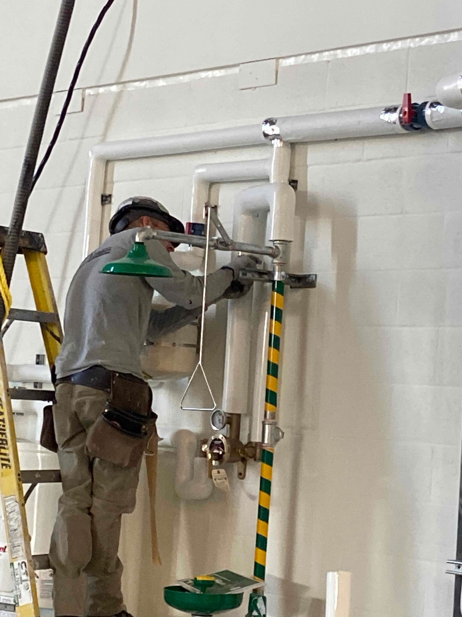 Un ouvrier portant un casque de chantier installe une douche de sécurité industrielle et un lave-yeux sur un mur carrelé de blanc.