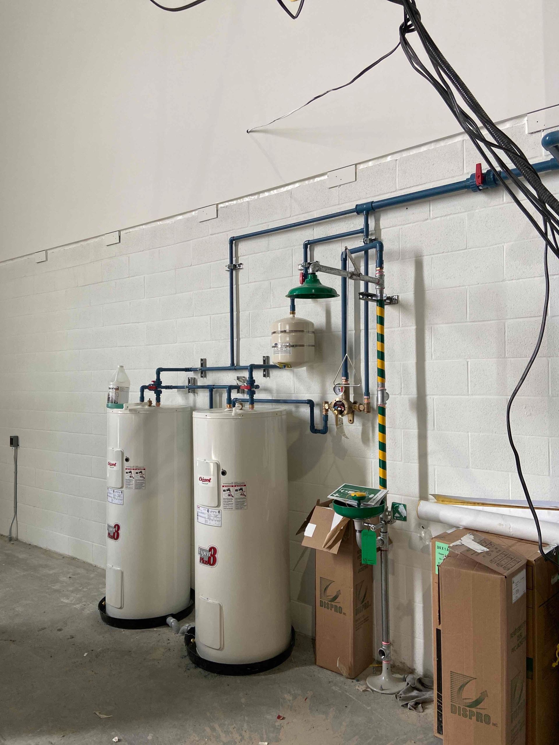 Deux chauffe-eau industriels avec tuyaux bleus et une douche de sécurité verte, fixés sur un mur de briques blanches.