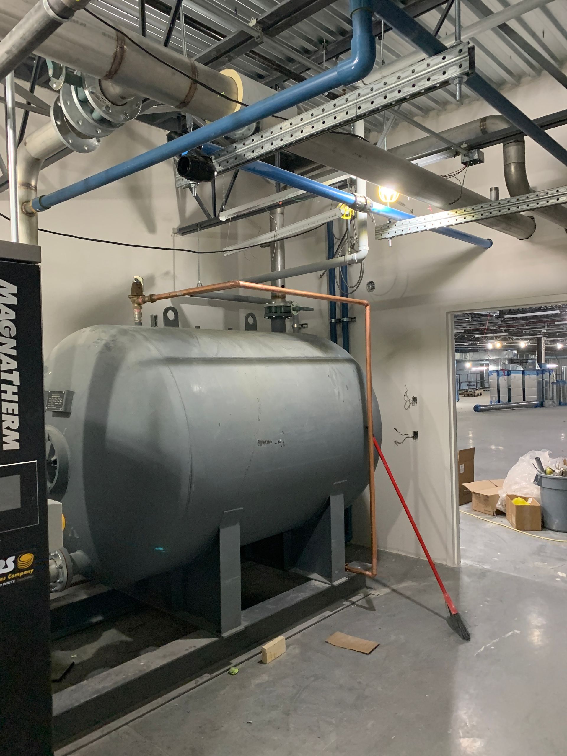 Dans une salle des machines, une grande cuve industrielle grise laisse apparaître des tuyaux au plafond, contre laquelle est appuyé un outil à manche rouge.