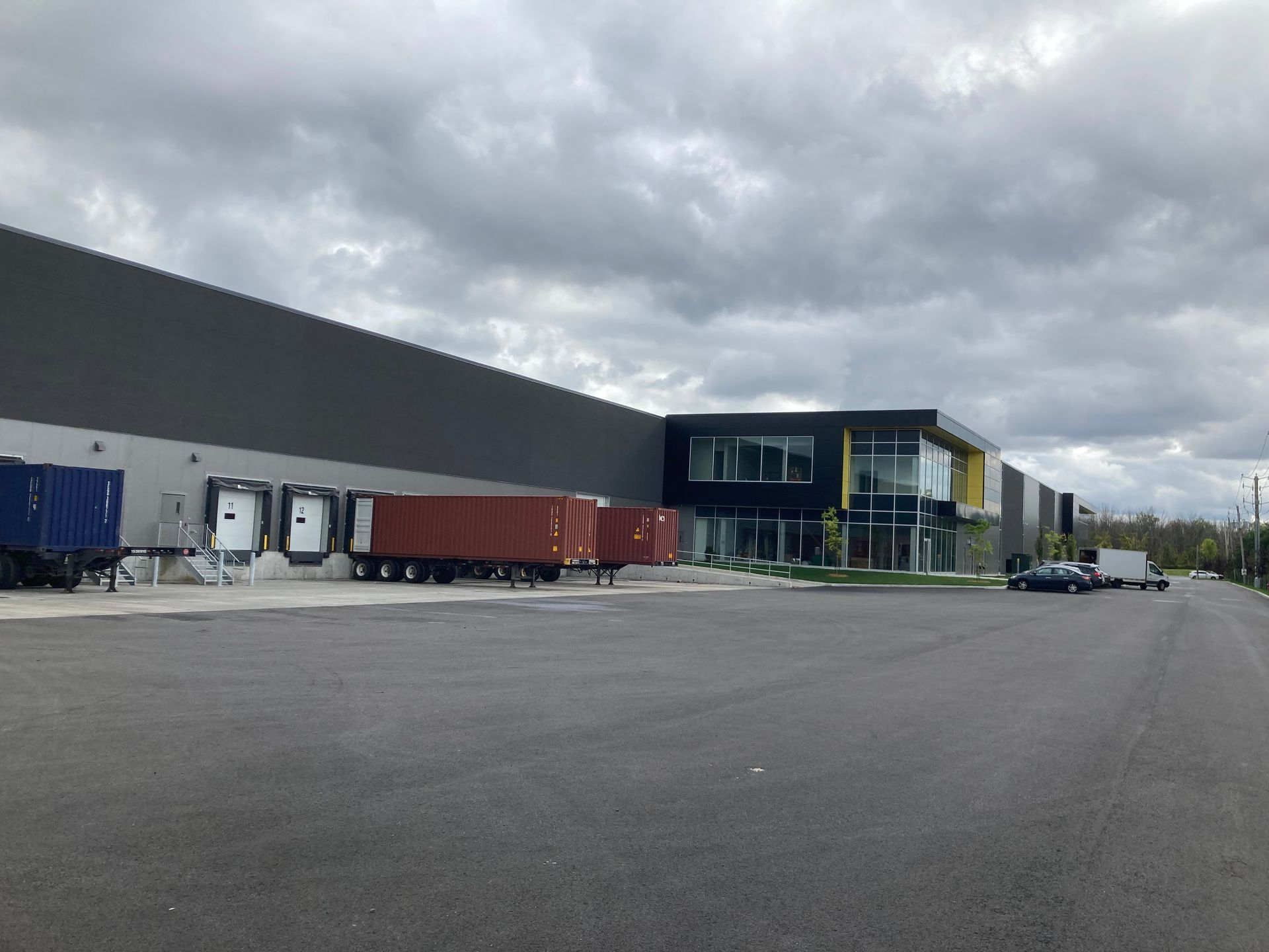 Un entrepôt moderne gris foncé avec des quais de chargement, des semi-remorques stationnées et des bureaux avec façade vitrée.