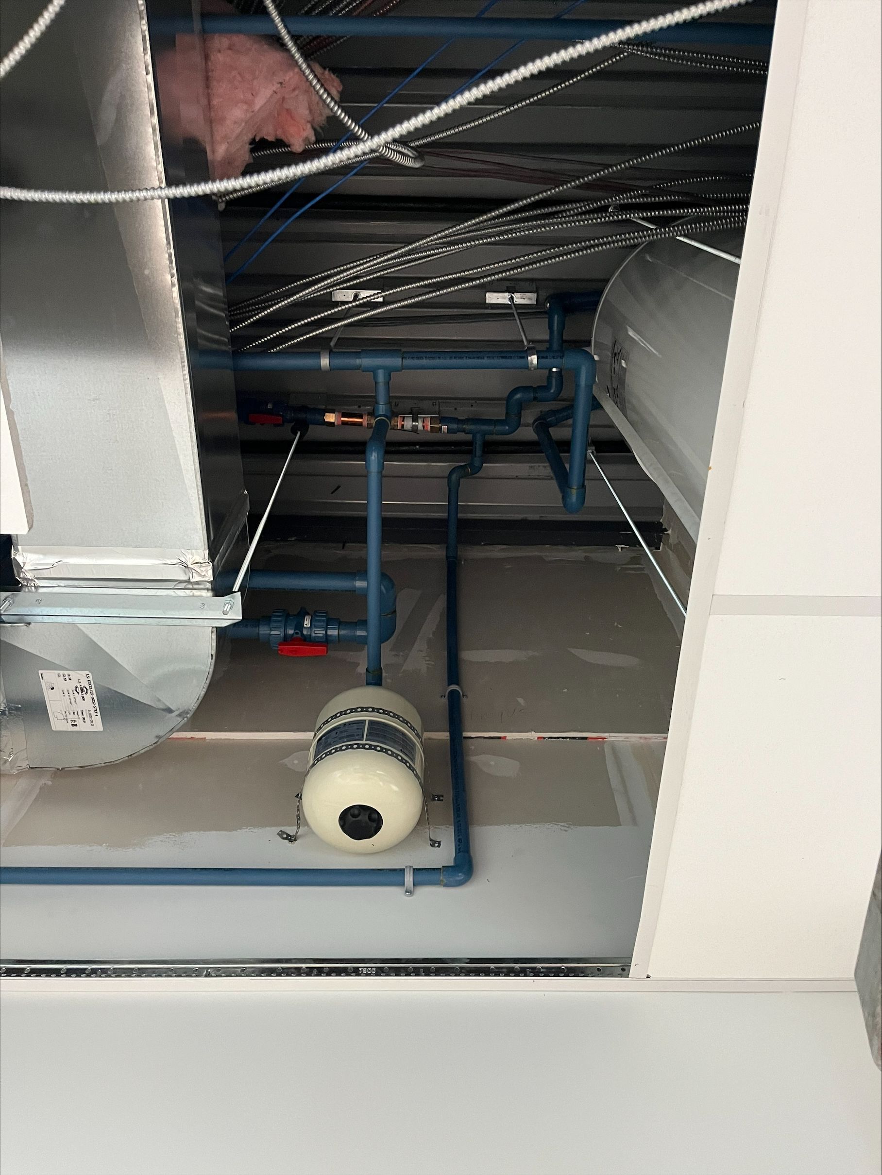 Espace plénum du plafond montrant des tuyaux bleus, des conduits métalliques, des câbles, de l'isolation et un vase d'expansion blanc.