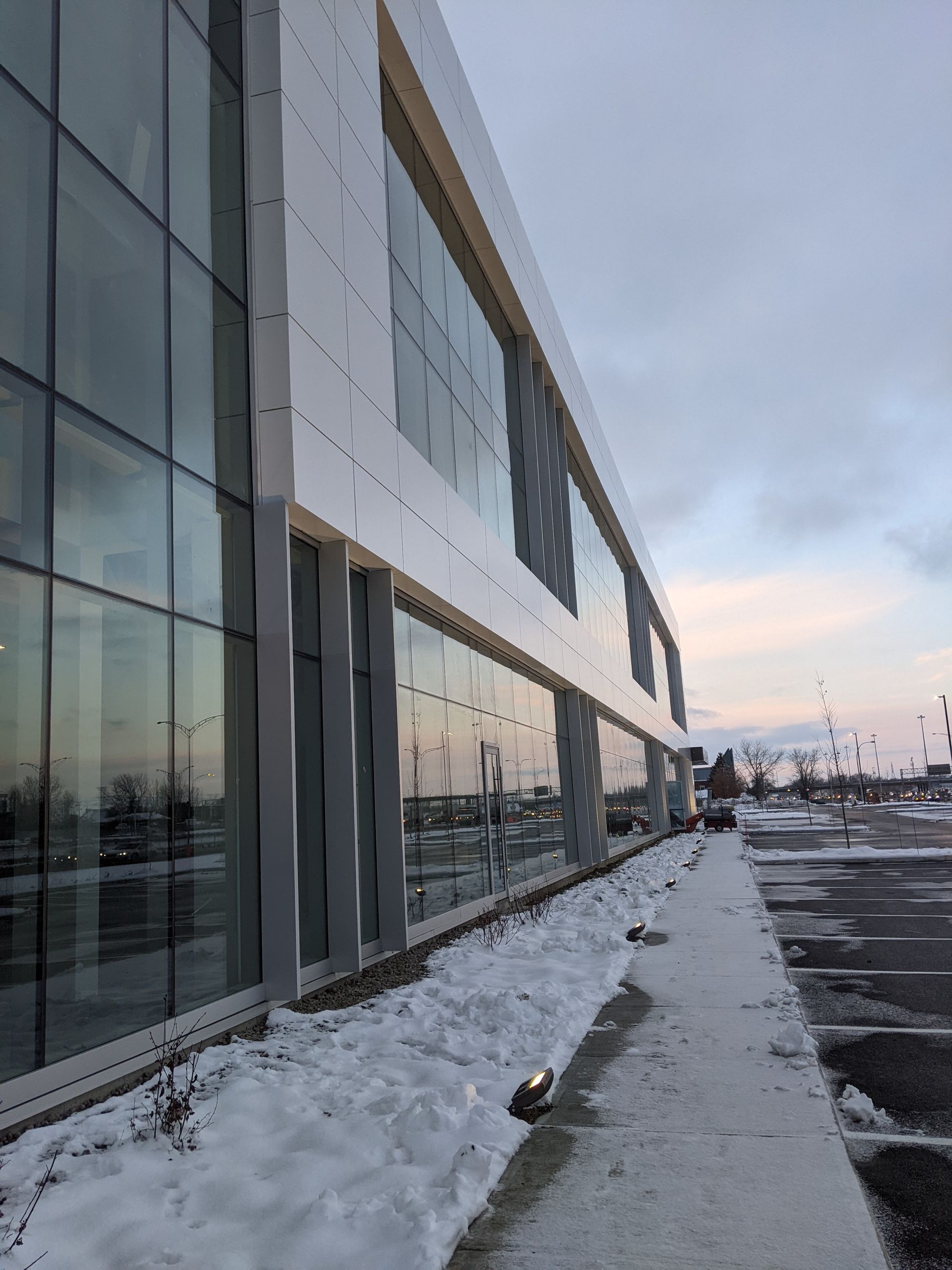 Un bâtiment moderne à plusieurs étages, doté d'une façade en verre et de panneaux réfléchissants, bordé d'une allée enneigée et d'un parking.