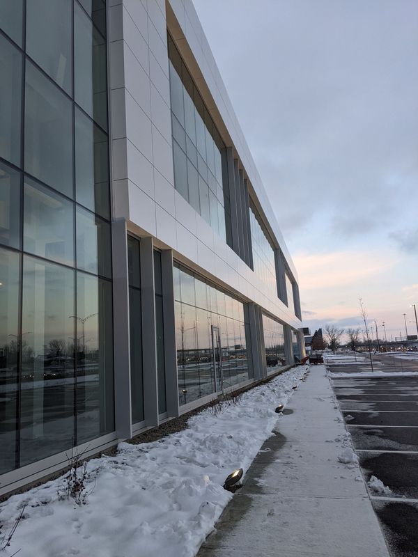 Un bâtiment moderne à plusieurs étages, doté d'une façade en verre et de panneaux réfléchissants, bordé d'une allée enneigée et d'un parking.