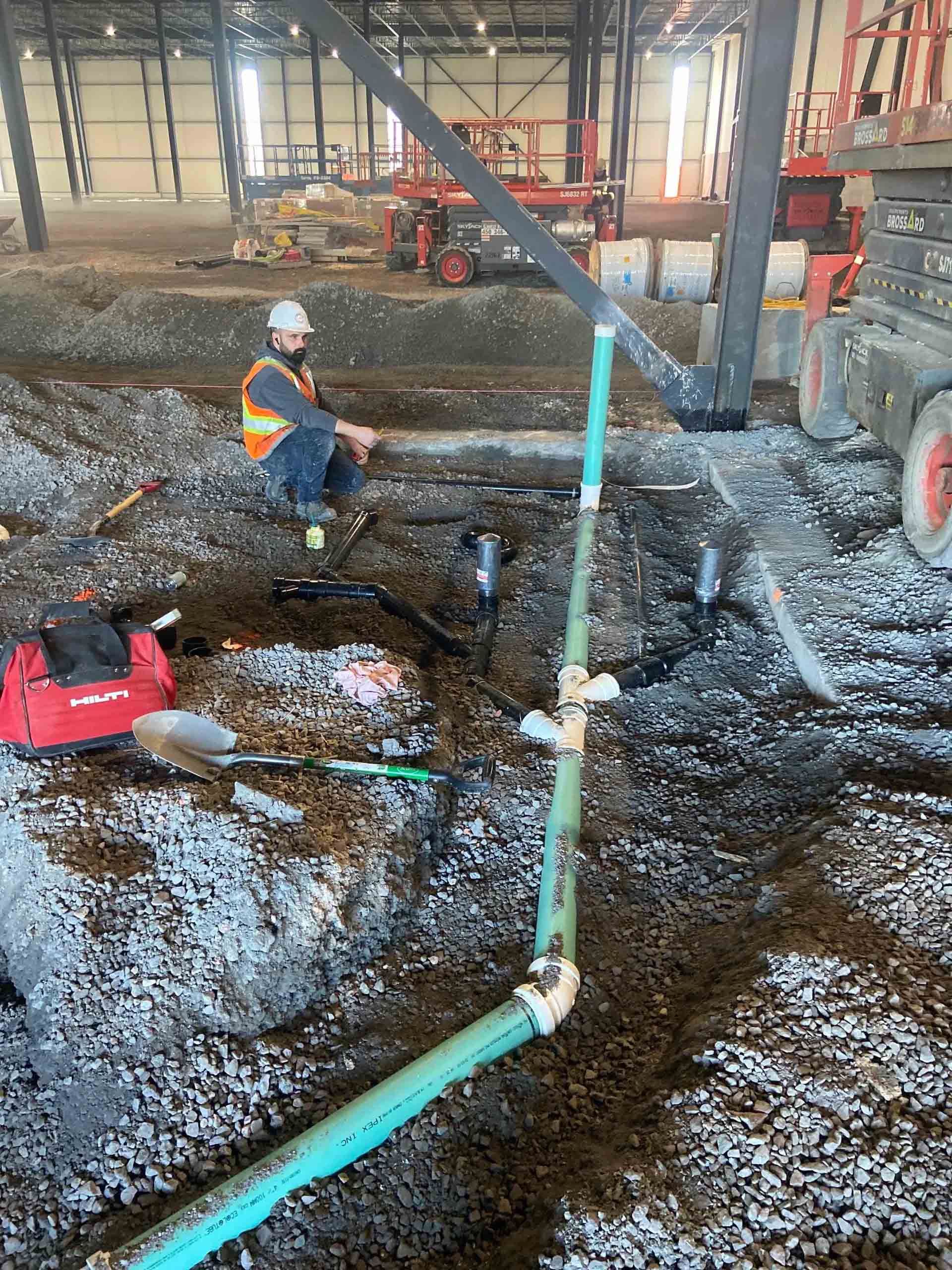 Un ouvrier portant un gilet de sécurité et un casque de chantier est agenouillé sur un chantier au sol de terre battue, près d'un tuyau vert et d'un sac rouge.