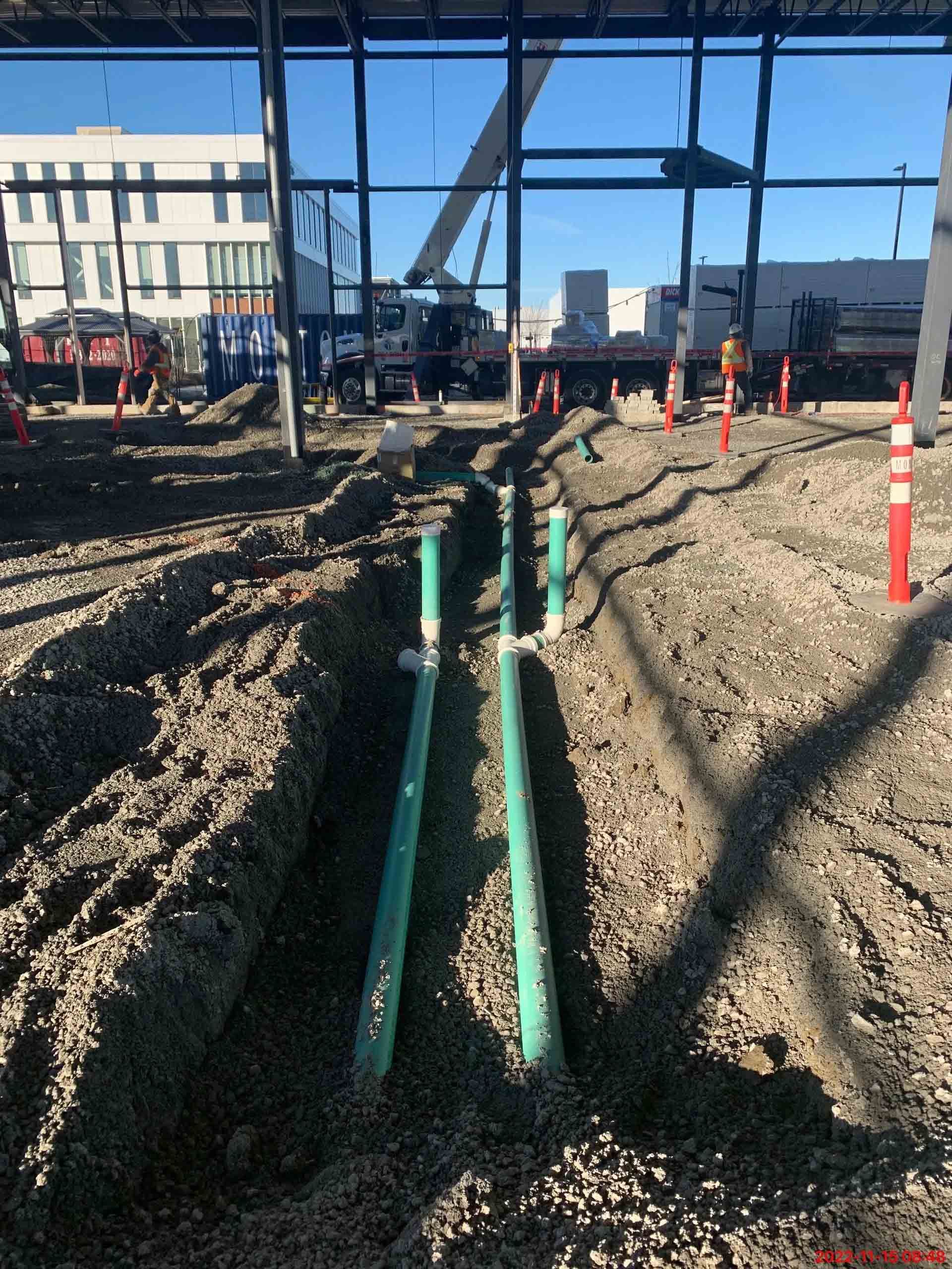 Deux tuyaux verts parallèles sont posés dans une tranchée de gravier sur un chantier de construction, avec une charpente métallique en arrière-plan.