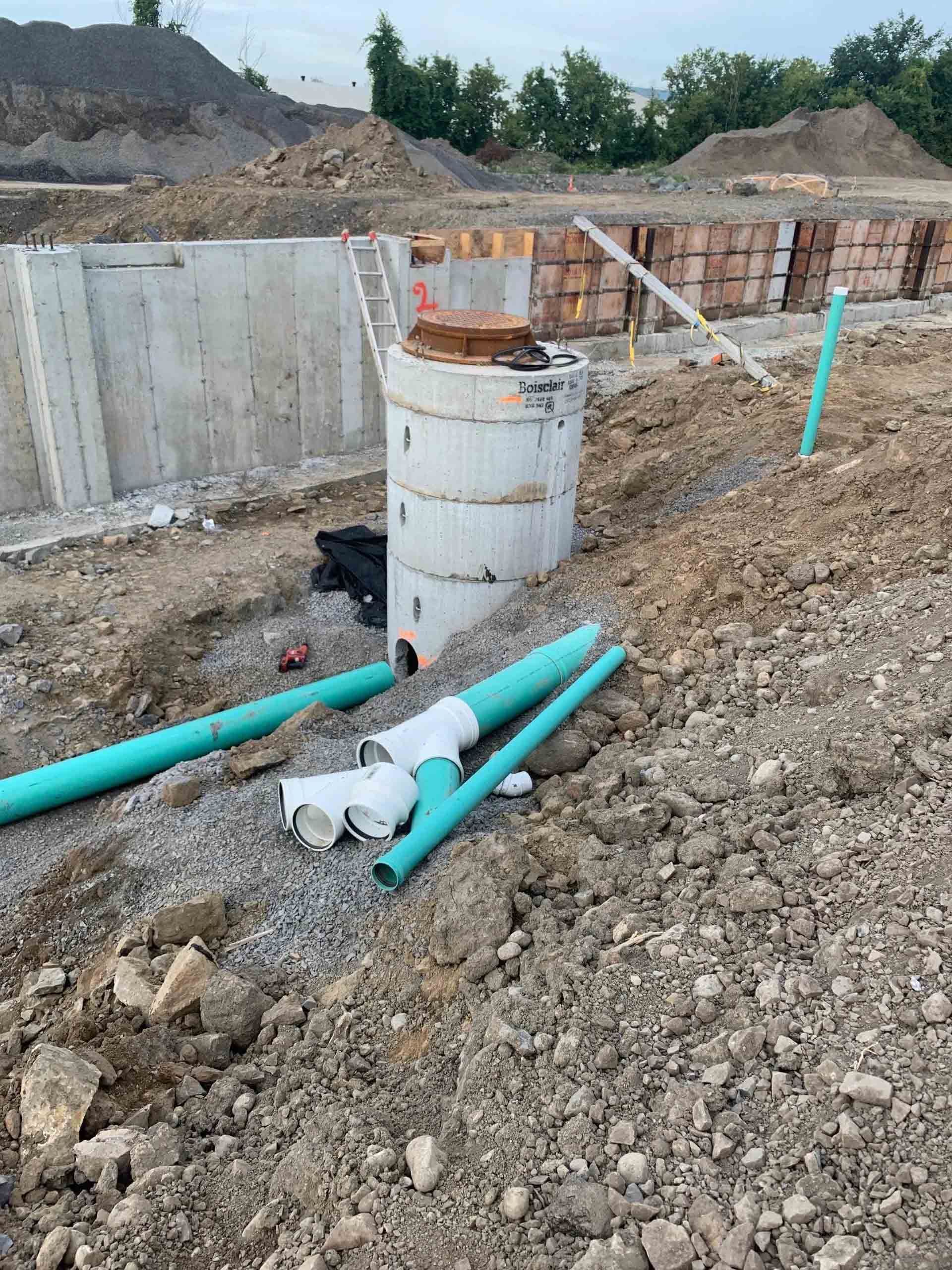 Chantier de construction avec une fosse en béton, un réservoir d'eau blanc et des tuyaux bleus sur un sol en gravier.