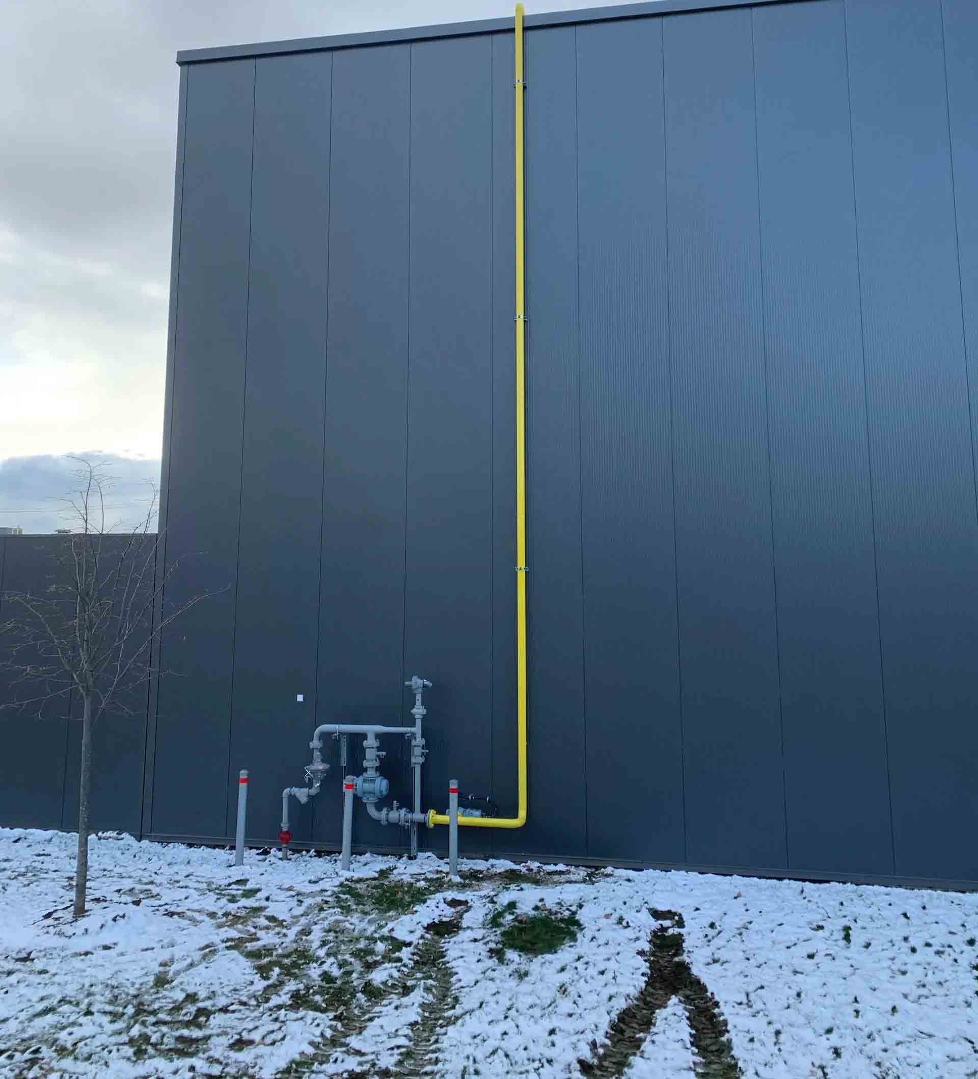 Un tuyau de gaz jaune court verticalement le long de la façade métallique sombre et ondulée d'un bâtiment, au-dessus de vannes de gaz extérieures grises.