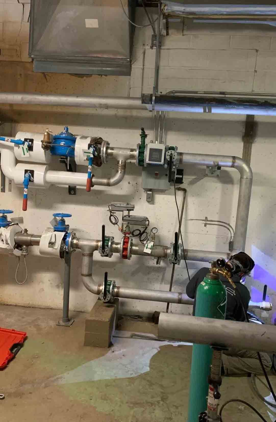 Un ouvrier en tenue de protection soude des tuyaux dans un local technique en sous-sol, comportant diverses vannes et des systèmes de plomberie métallique.