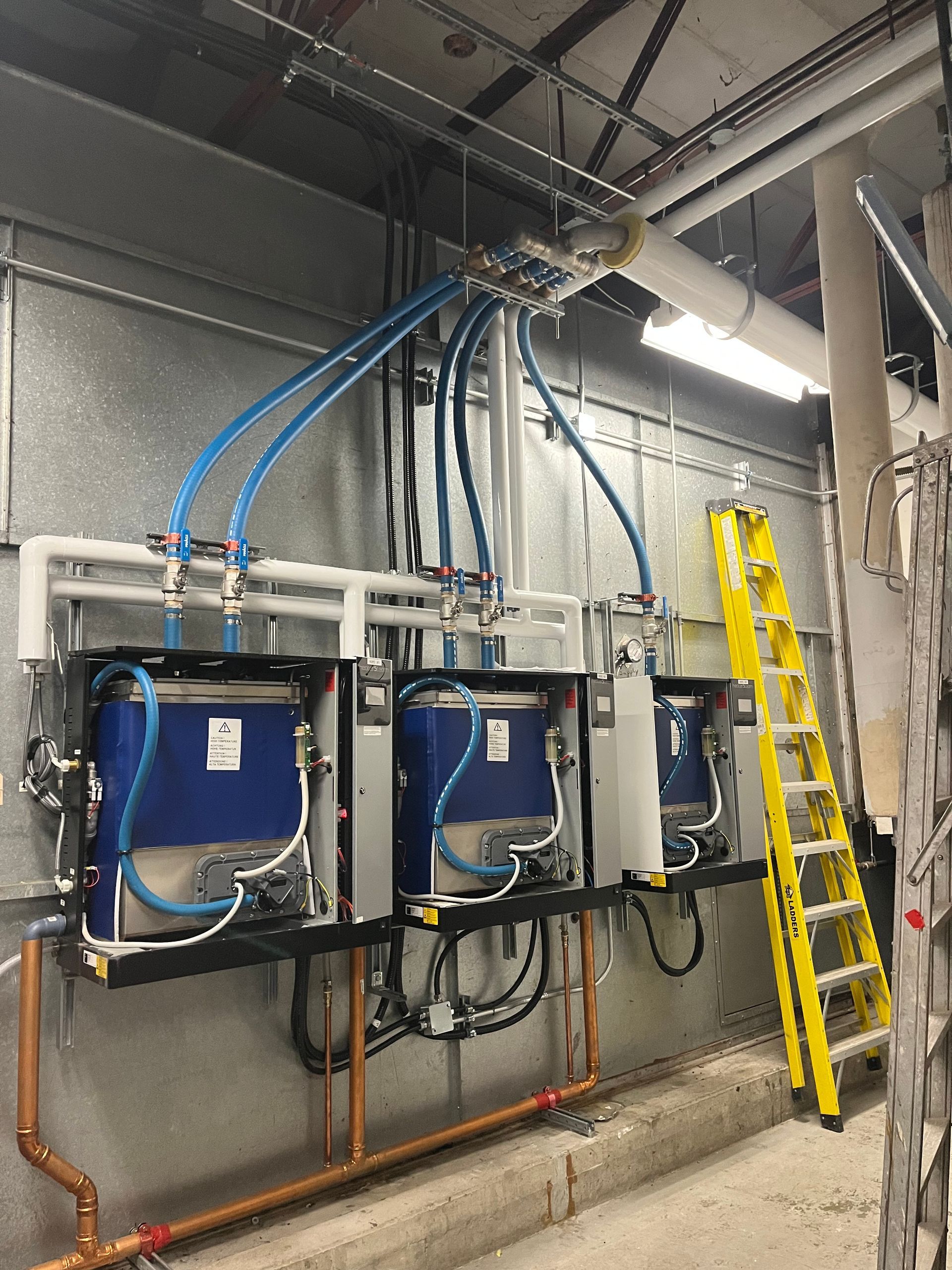 Trois chauffe-eau bleus fixés au mur, avec des tuyaux blancs et des flexibles bleus, dans un environnement industriel, avec une échelle jaune.