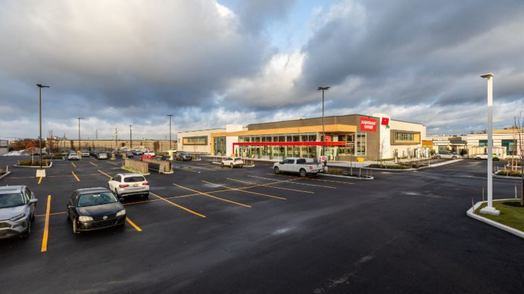 Un bâtiment commercial moderne avec une enseigne rouge, vu depuis un parking par temps nuageux, avec des voitures garées aux emplacements marqués.