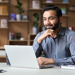 Man smiling at a laptop in a cozy office, resting his chin on his hand