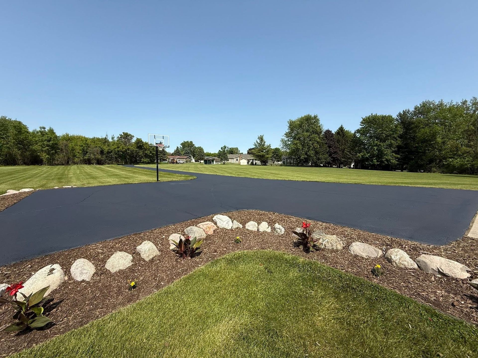 Curved blacktop driveway under a blue sky, bordered by rocks and grass in a landscaped yard