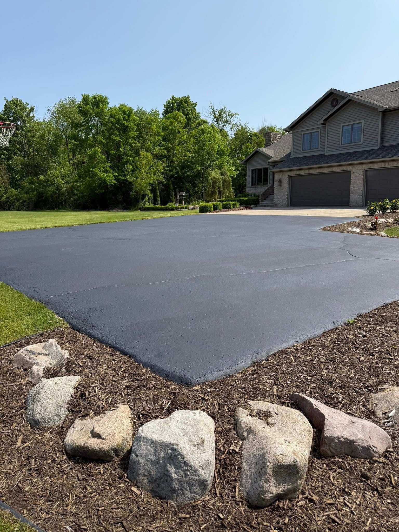 Freshly paved dark driveway beside a suburban house, bordered by rocks and trees