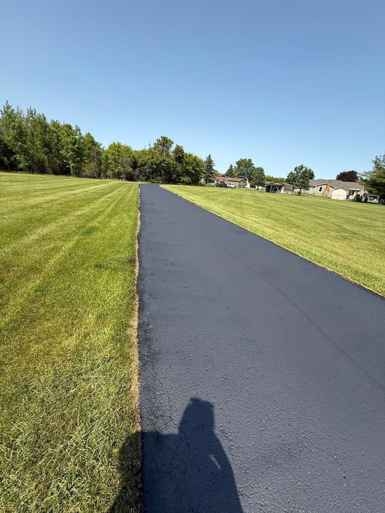 Paved path through a grassy park under a clear blue sky, with a shadow in the foreground.