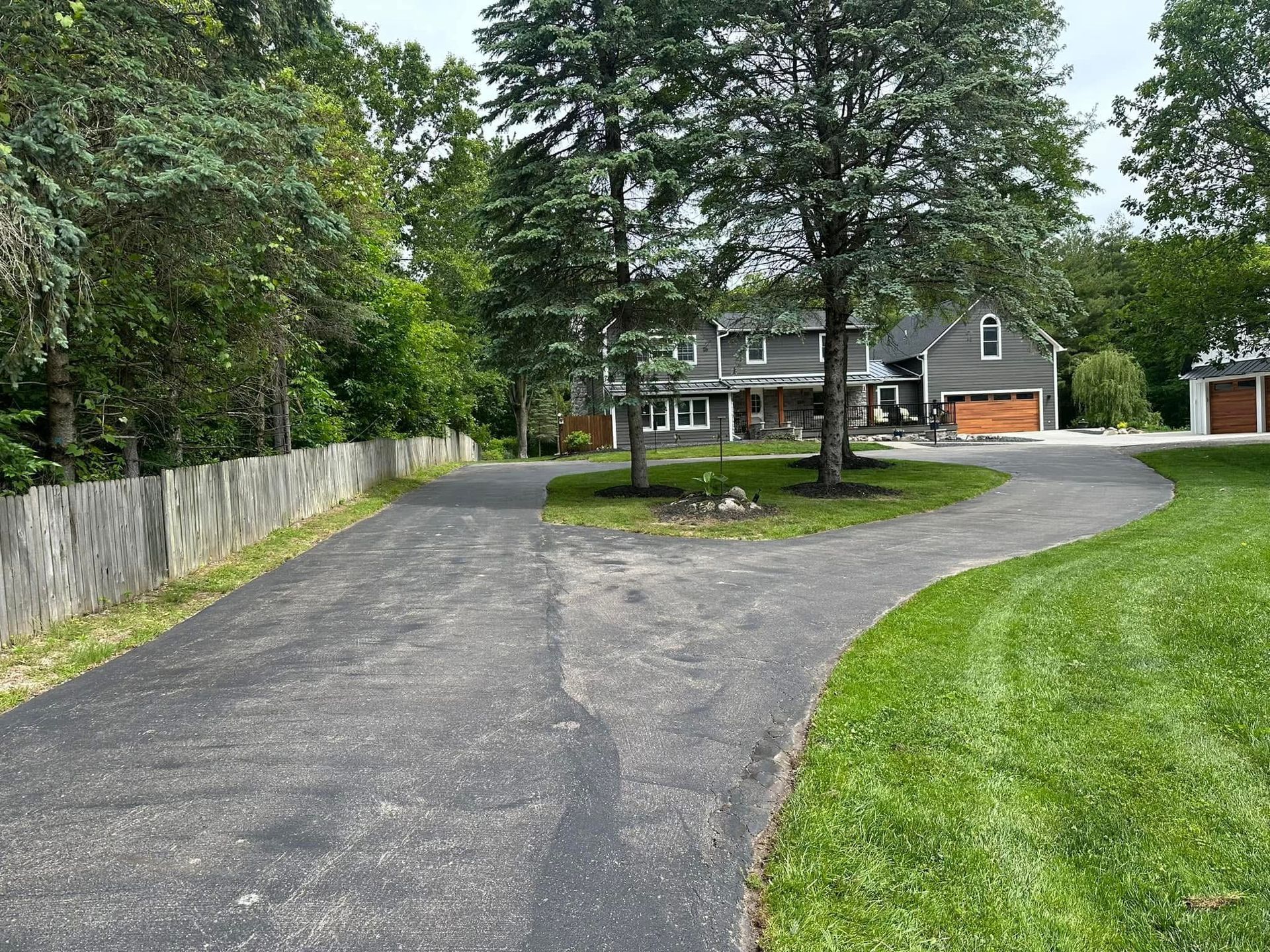 Curved driveway leading to a gray house with trees and a green lawn.