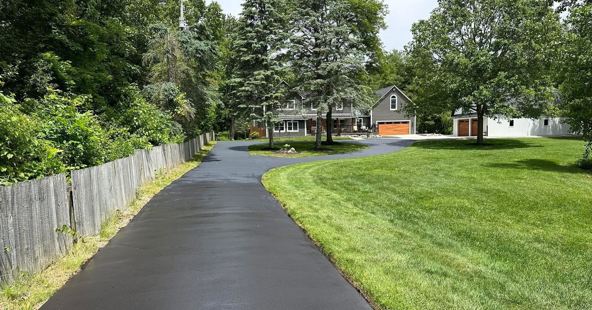 Curving black paved path beside a wooden fence and green lawn, leading to houses in a sunny park-like area