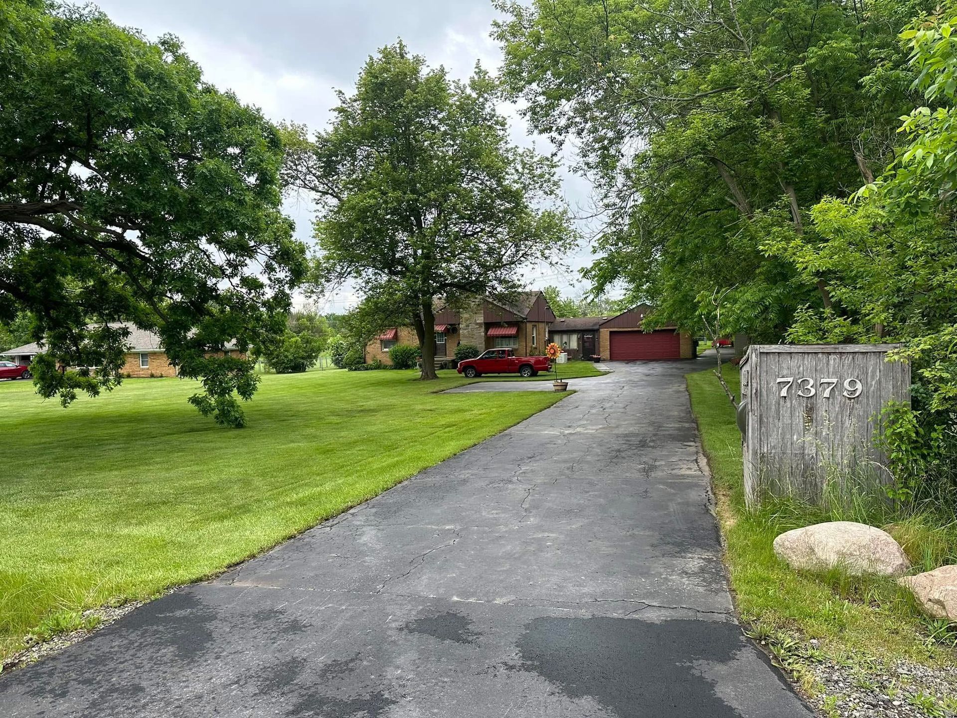 Driveway leading to a red house with garage, bordered by green lawn and trees, with a stone address marker.