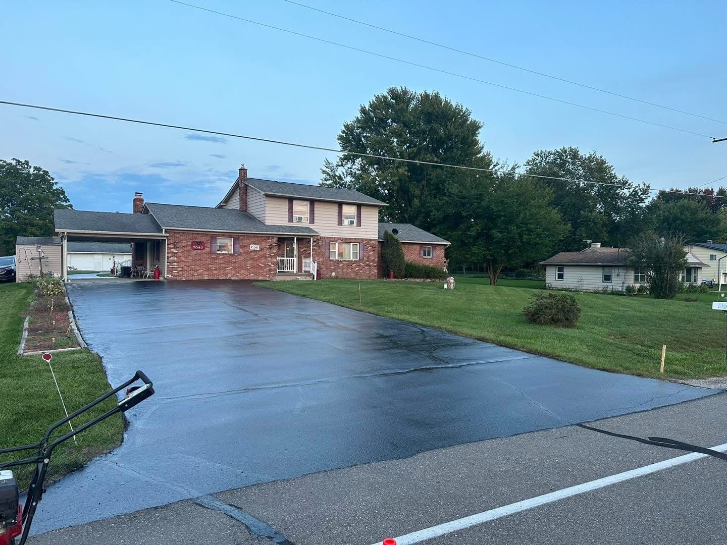 Wet suburban house with driveway, lawn, and large tree under a blue sky