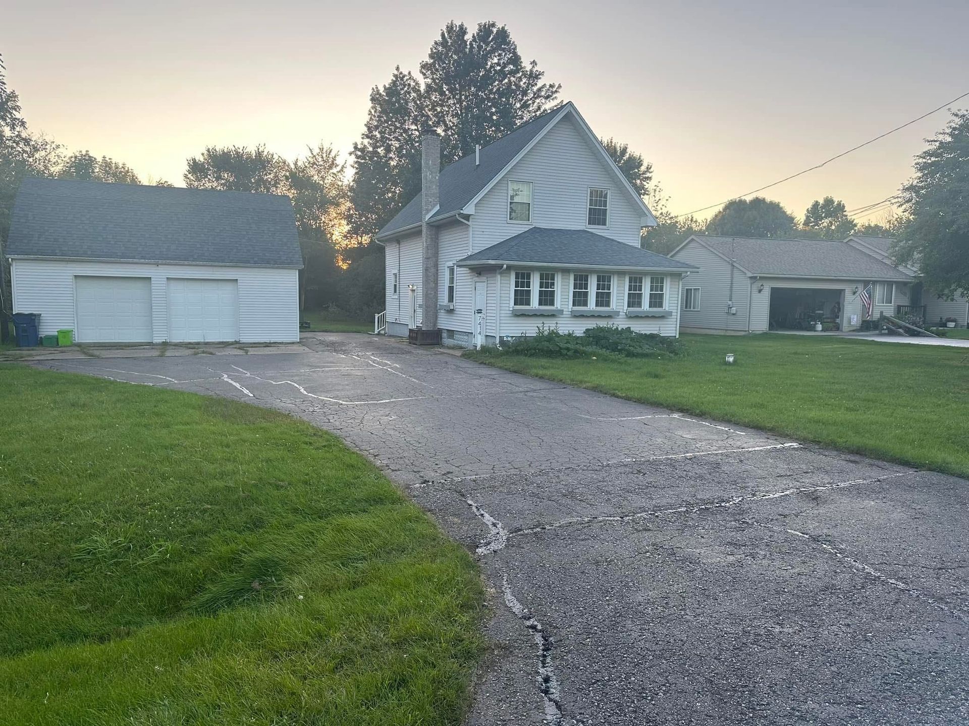 Gravel driveway leading to a white house and detached garage at dawn, with green lawn and trees.