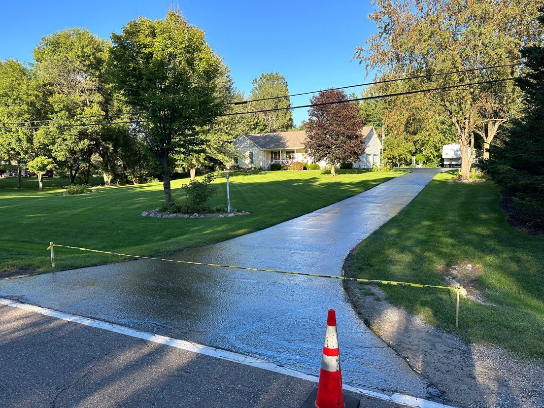 Freshly paved driveway beside a grassy yard with trees and houses, marked by orange cones