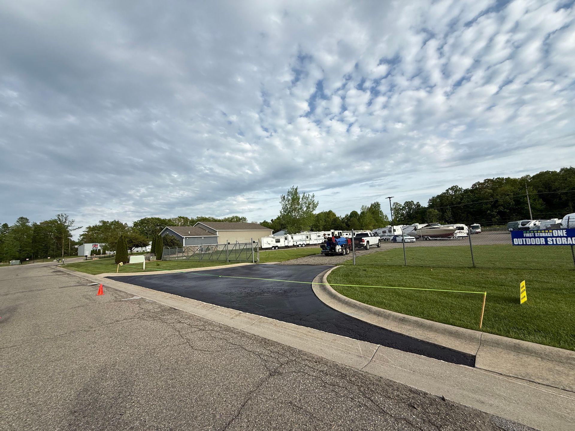 Curved parking lot lane beside a grassy field under a cloudy sky, with parked vehicles and cones in the distance