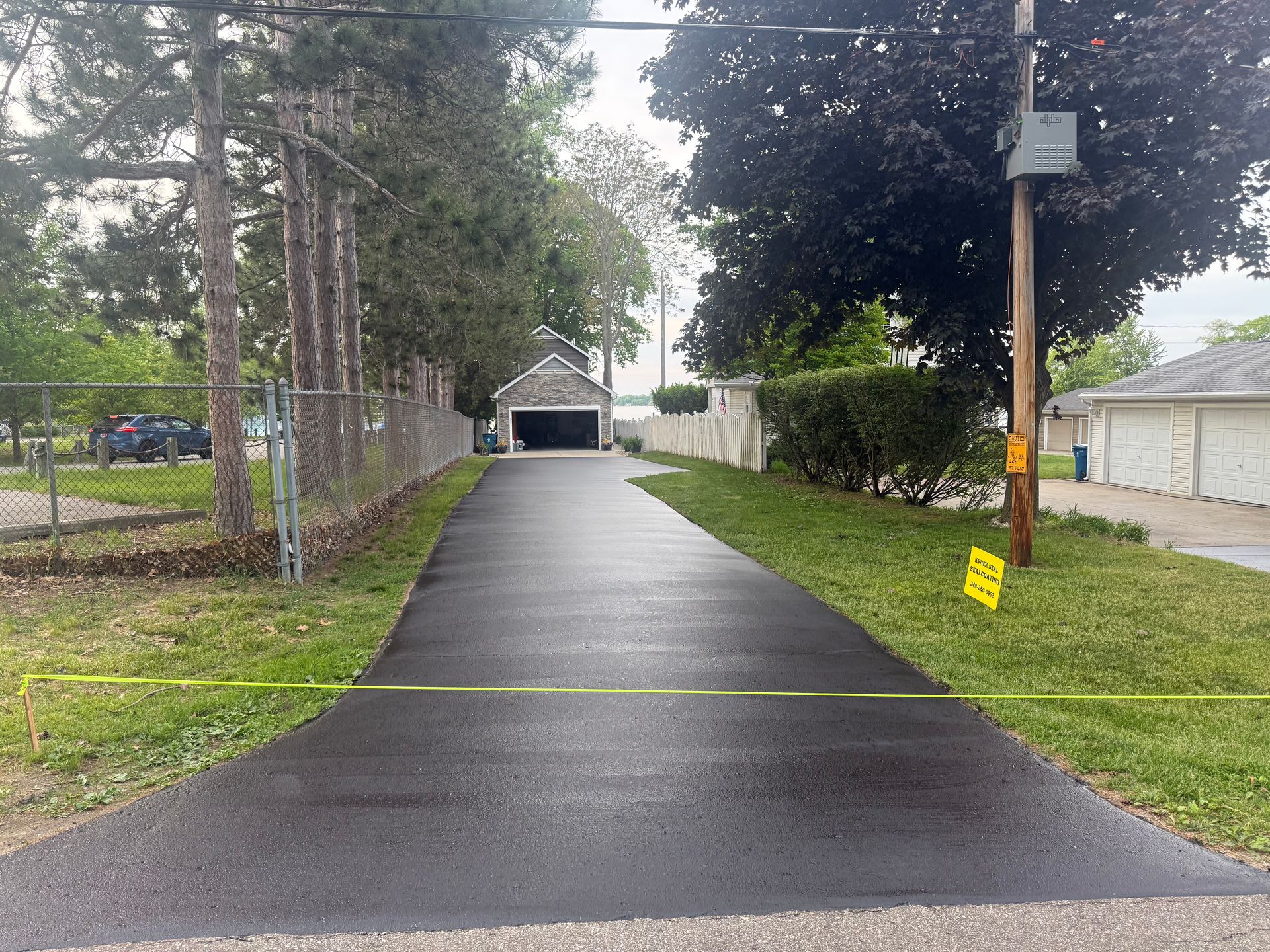 Residential driveway with fresh black asphalt leading to a garage, flanked by trees and a utility pole.