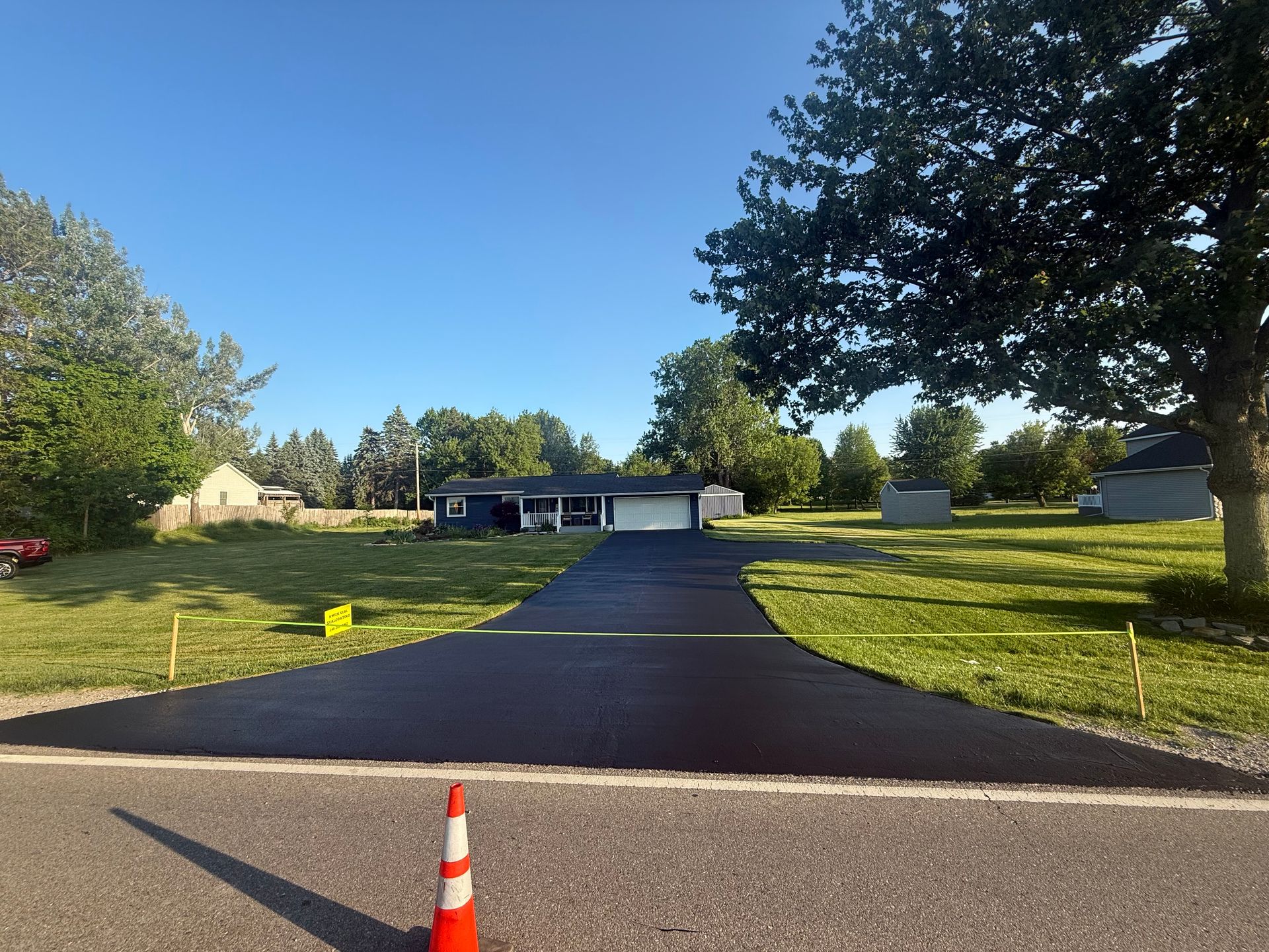 Long black driveway leading to a house with a garage, bordered by green grass and trees under a blue sky