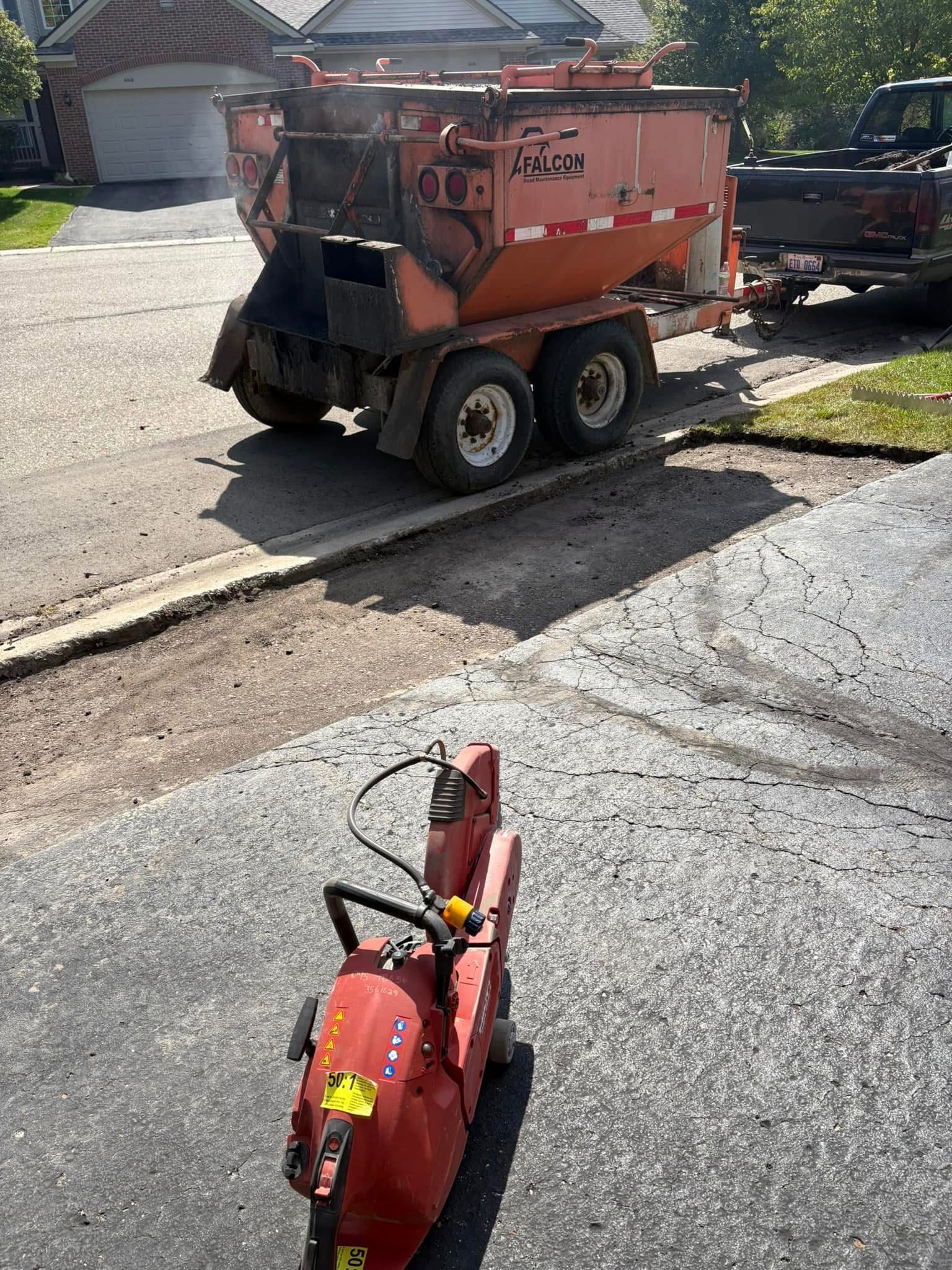 Orange stump grinder parked on a driveway, with a red snowblower in the foreground and houses nearby.