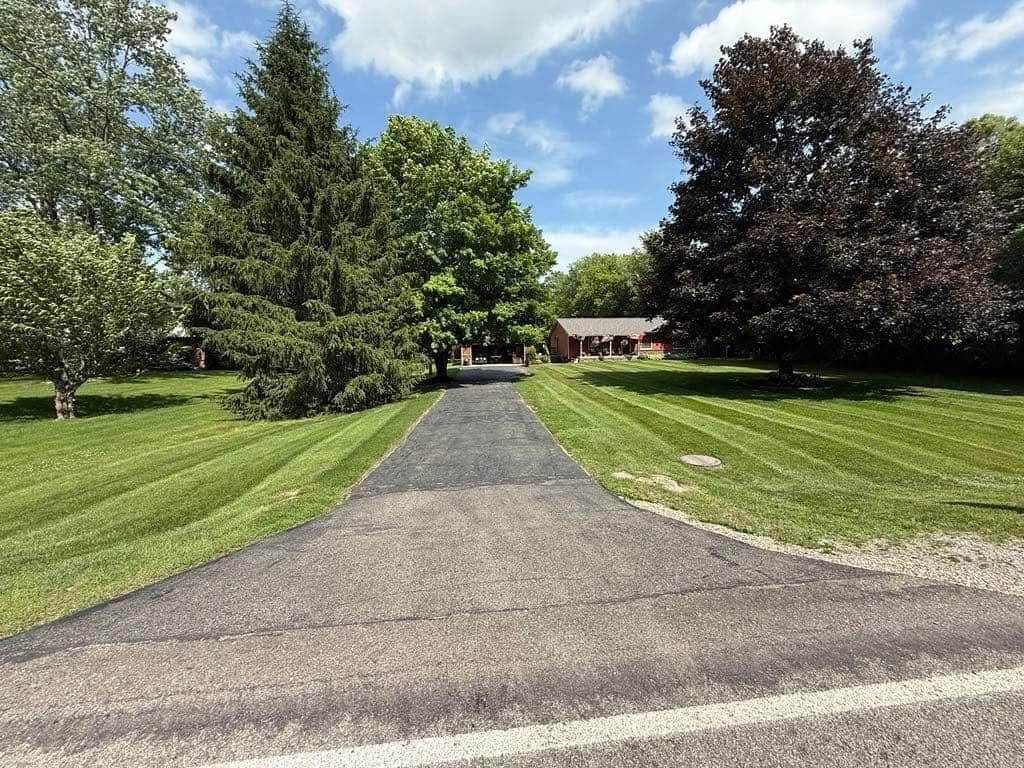 Paved driveway leading to a house, lined with green lawns and tall trees under a blue sky.
