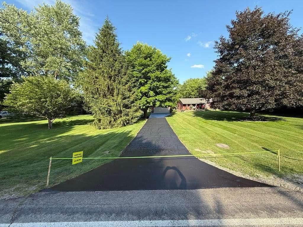 Gravel driveway leading to a house, bordered by green lawns and trees under a blue sky