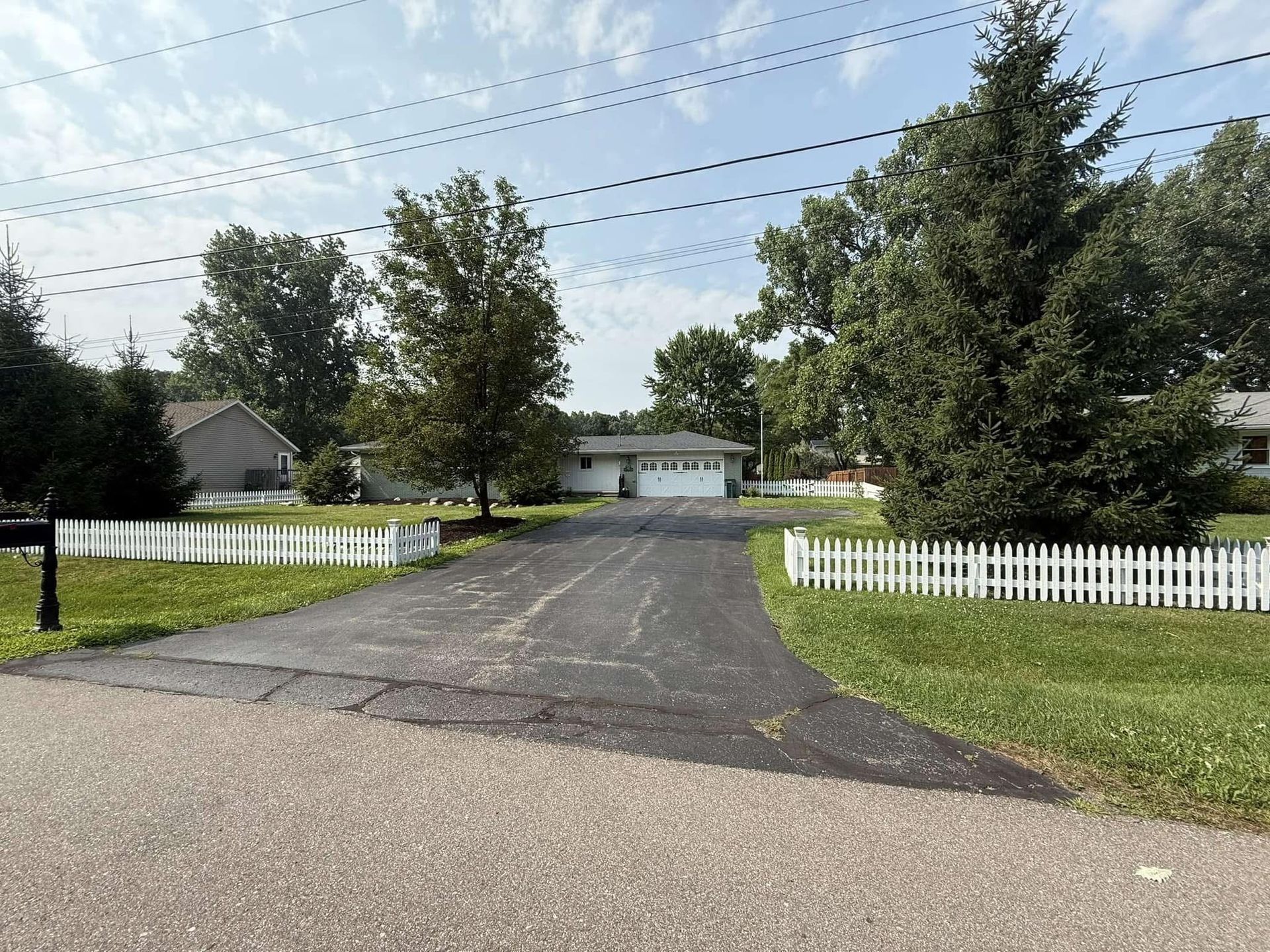 Residential driveway with white picket fences, trees, and houses under a cloudy sky