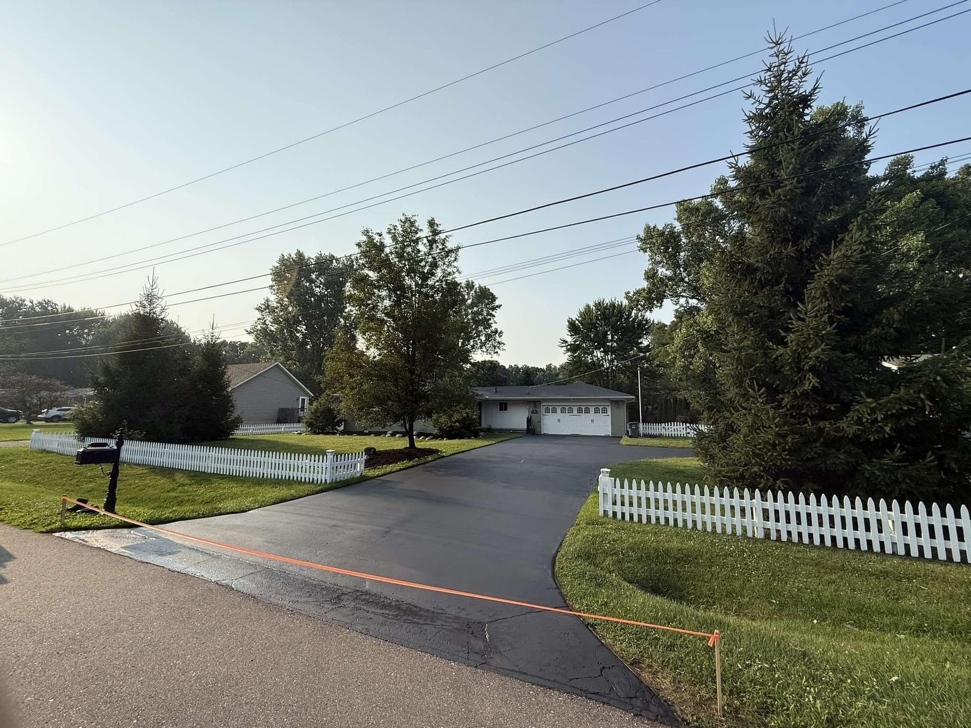 Driveway leading to a garage, with white picket fence, trees, and wet pavement at dusk.