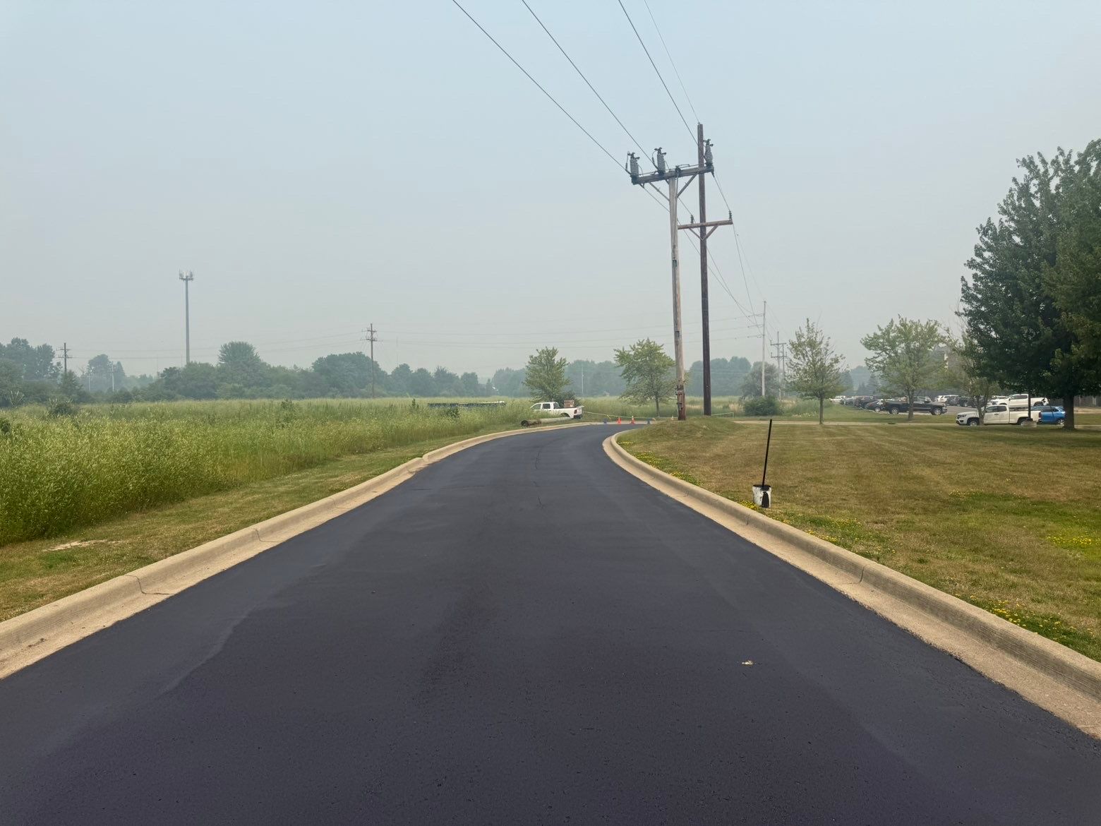 Curving paved road beside grassy fields and utility poles under a hazy sky