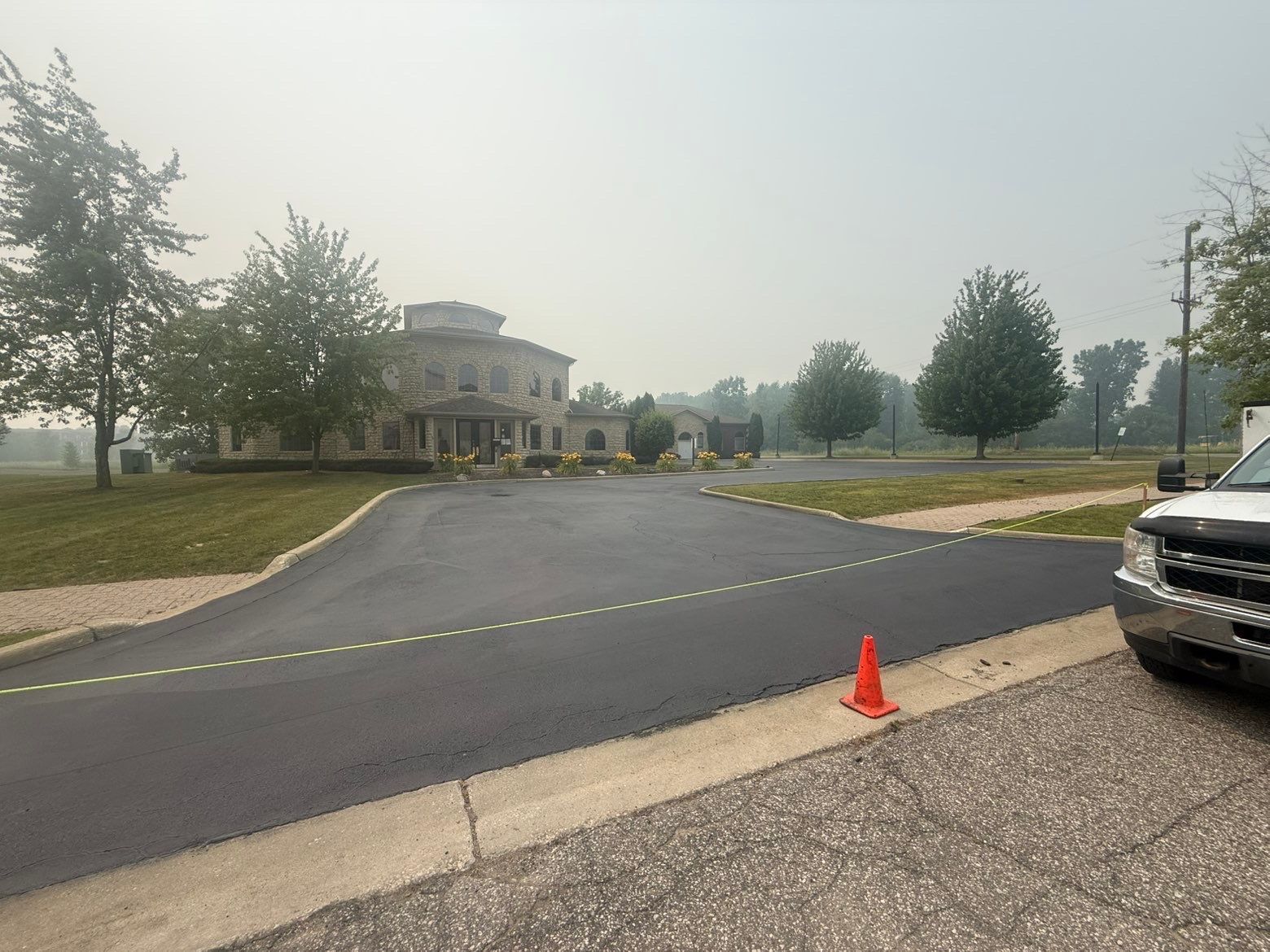 Paved driveway beside a large building, marked with yellow tape and an orange cone in a hazy outdoor setting