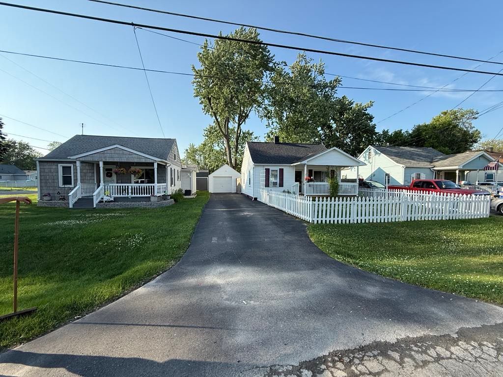 Two small houses with a driveway between them, one with a white picket fence, on a sunny street.