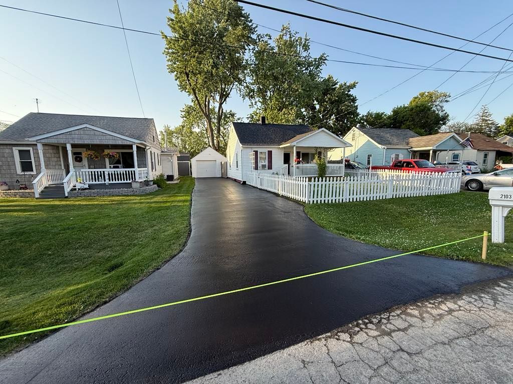 Quiet suburban street with a small white house, driveway, lawns, and a chain-link fence under a bright sky.