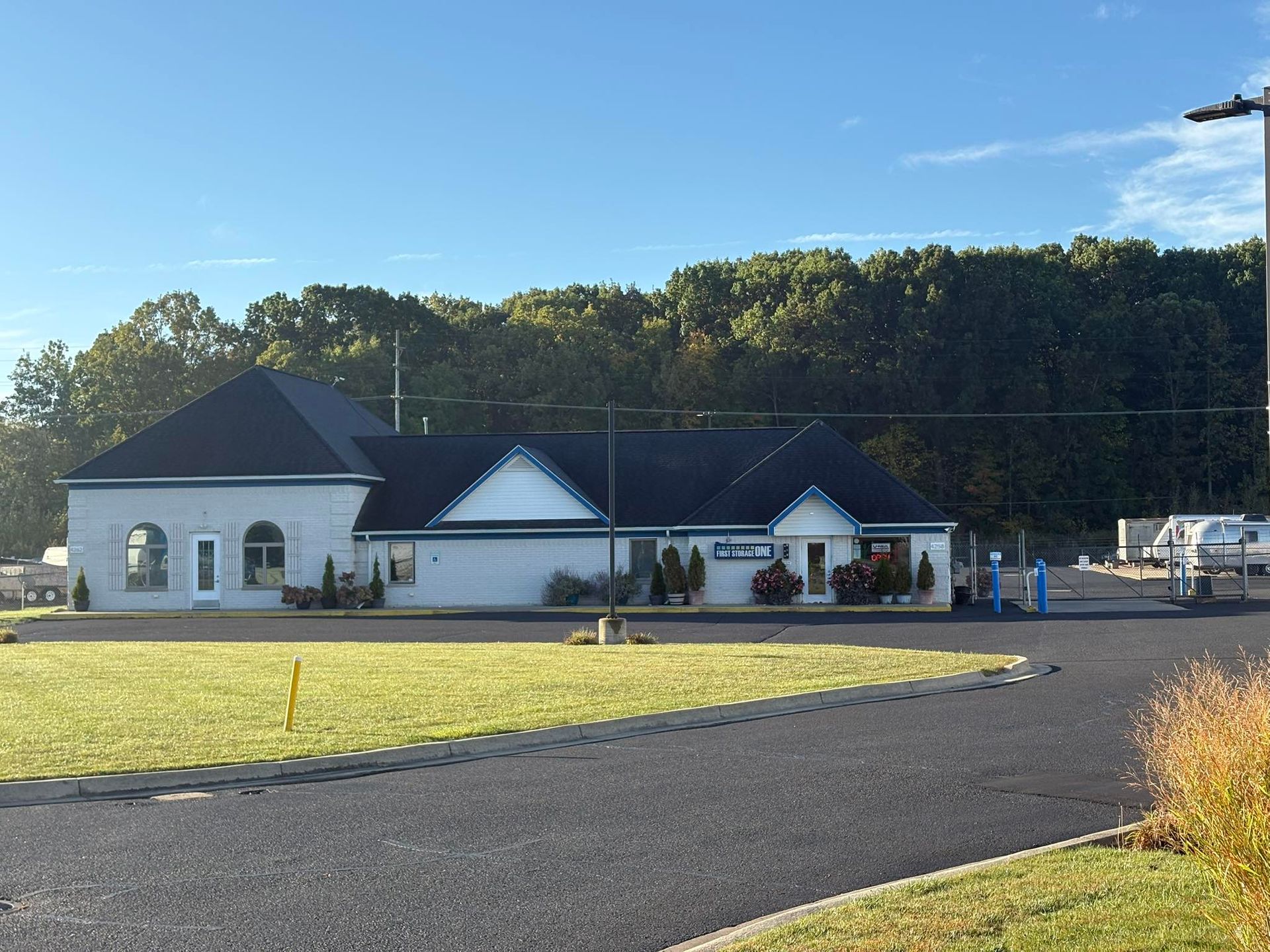 White building with dark roof beside a gravel lot and grassy field, with people gathered near the entrance.