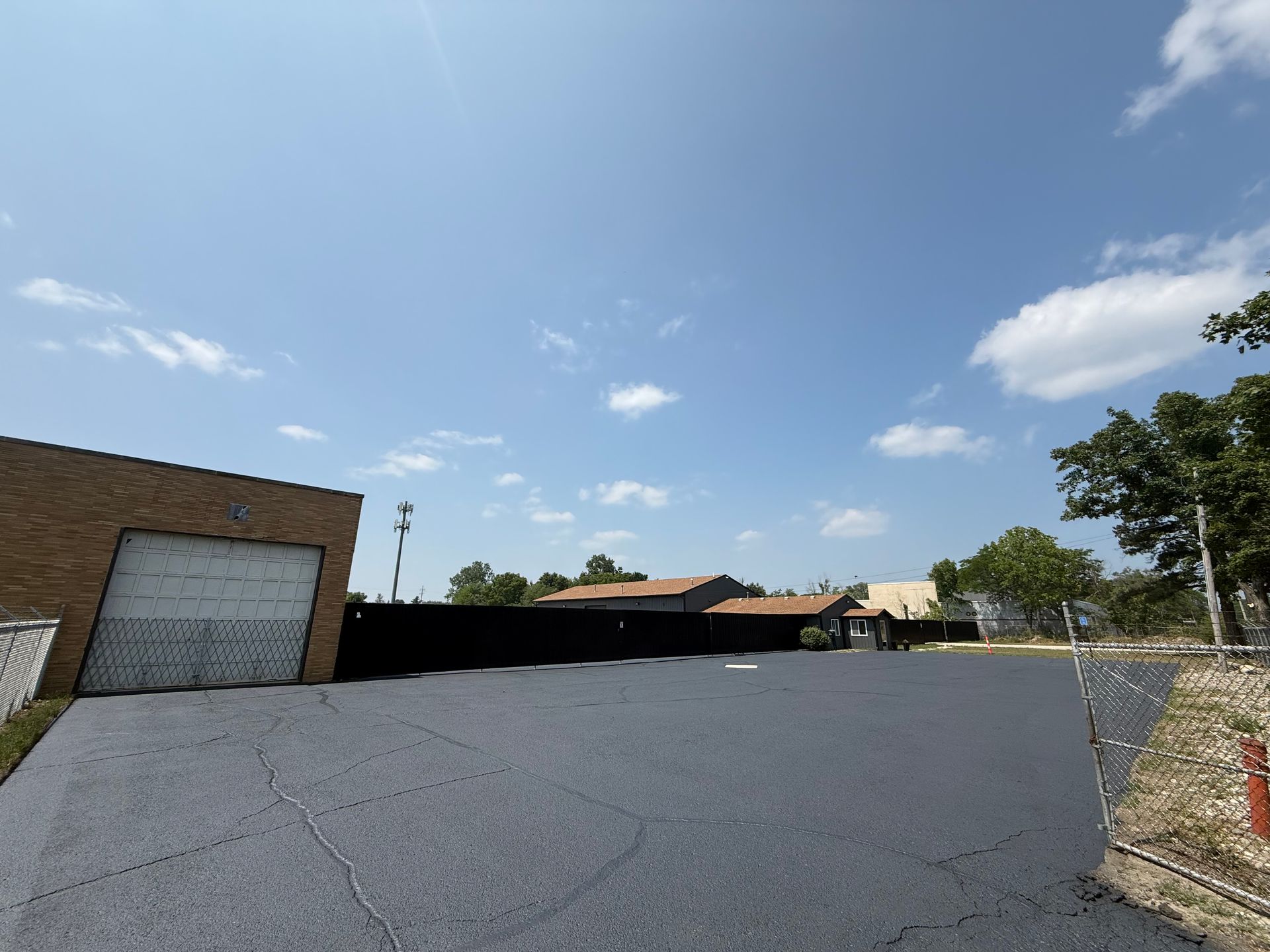 Empty paved lot beside brick buildings under a blue sky, with trees and a black fence in the distance