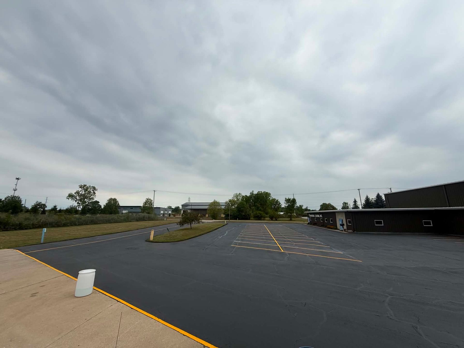 Empty parking lot under cloudy sky, with a few cars and trees in the distance.