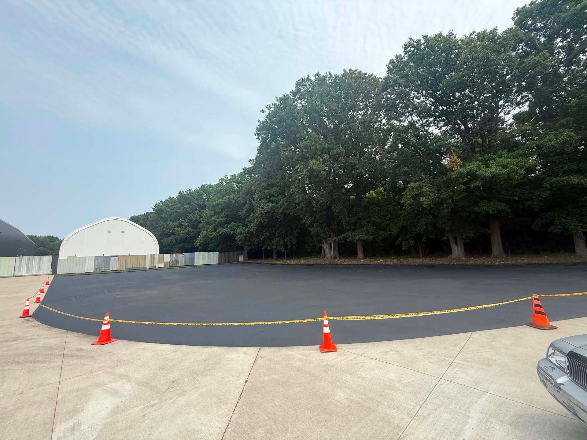 Empty paved lot with orange cones, yellow caution tape, trees, and a white tent in the background