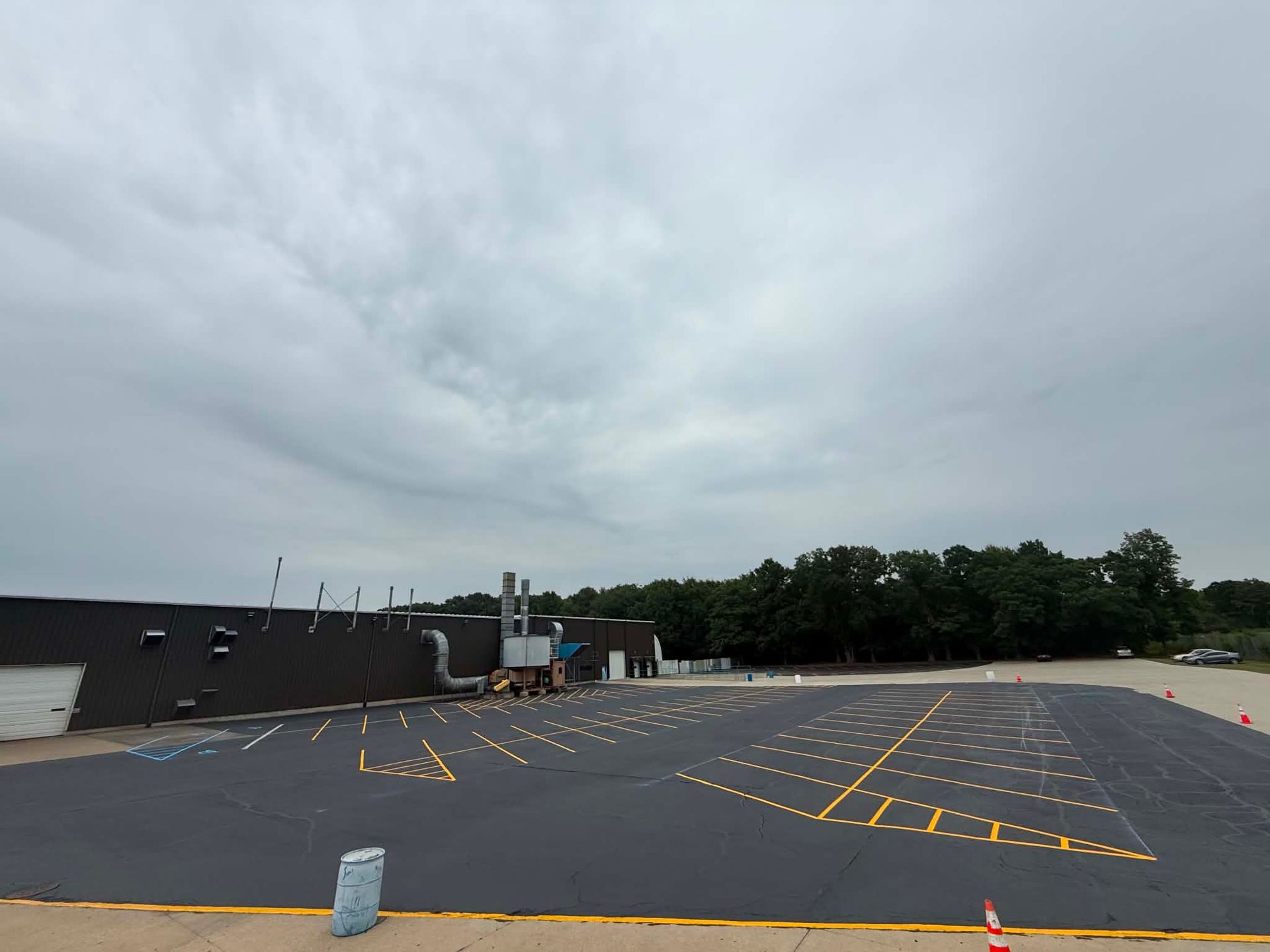 Empty parking lot with yellow painted lines under a cloudy sky, beside a low building and trees