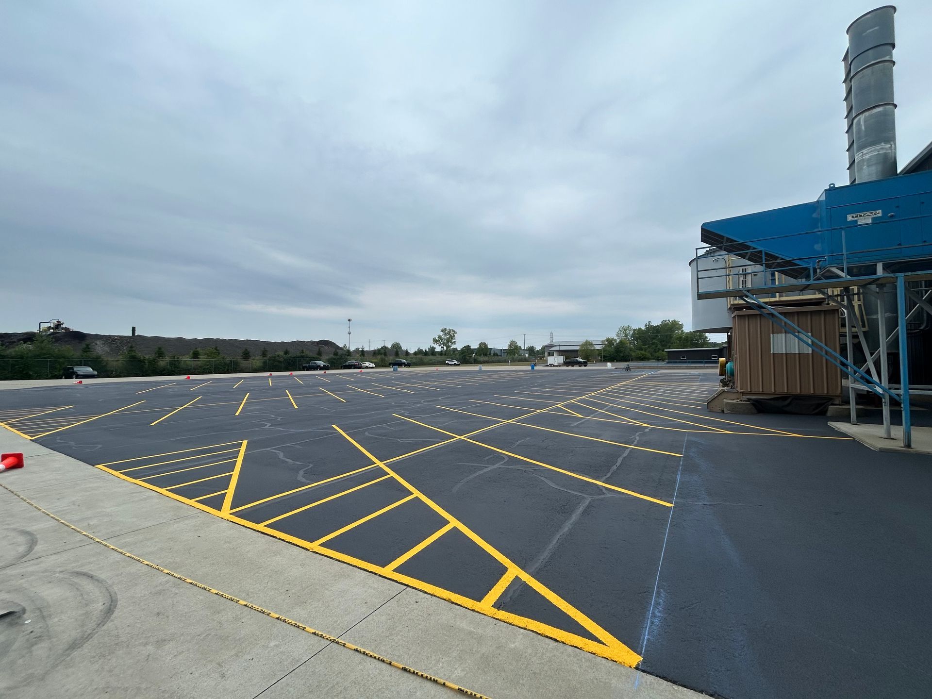 Empty paved lot with yellow diagonal markings beside an industrial building under a cloudy sky