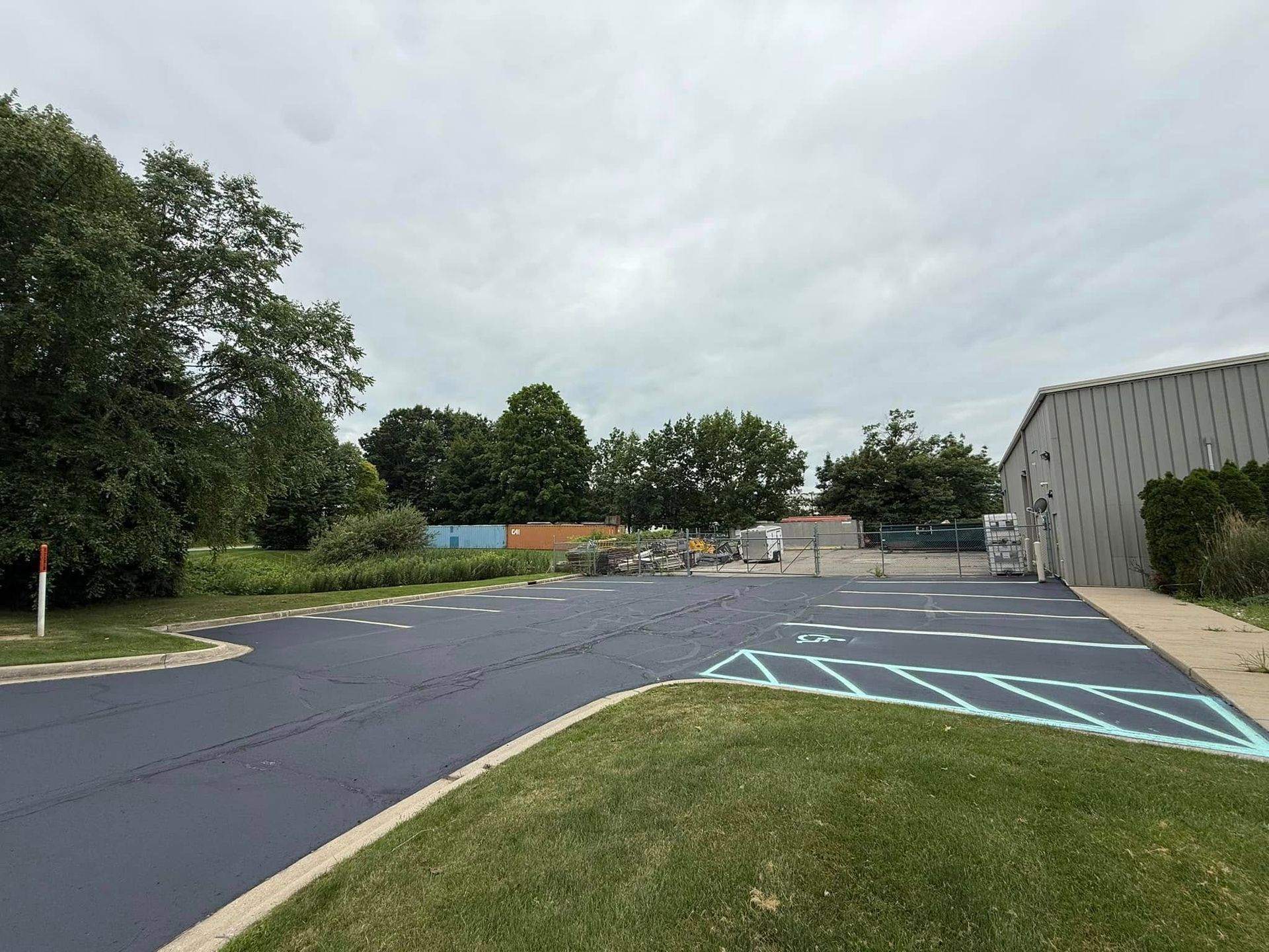 Empty parking lot beside a gray building, with blue-striped accessible parking spaces and trees under cloudy skies